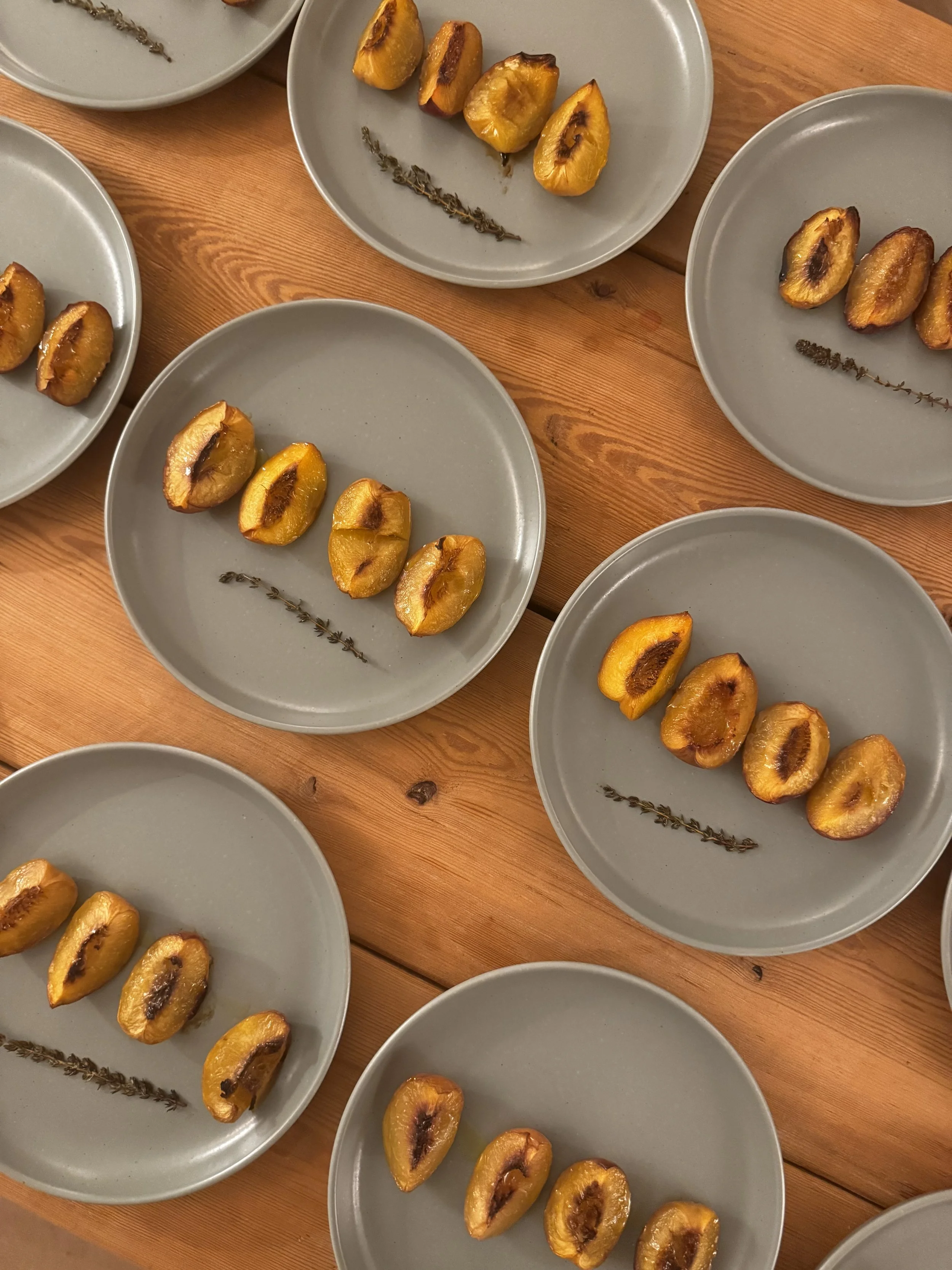 Multiple gray plates with three halved yellow peaches each, arranged in a row on a wooden table, some with visible brown spots and a sprig of herbs. Nourishing yoga retreat food in Devon and Portugal.
