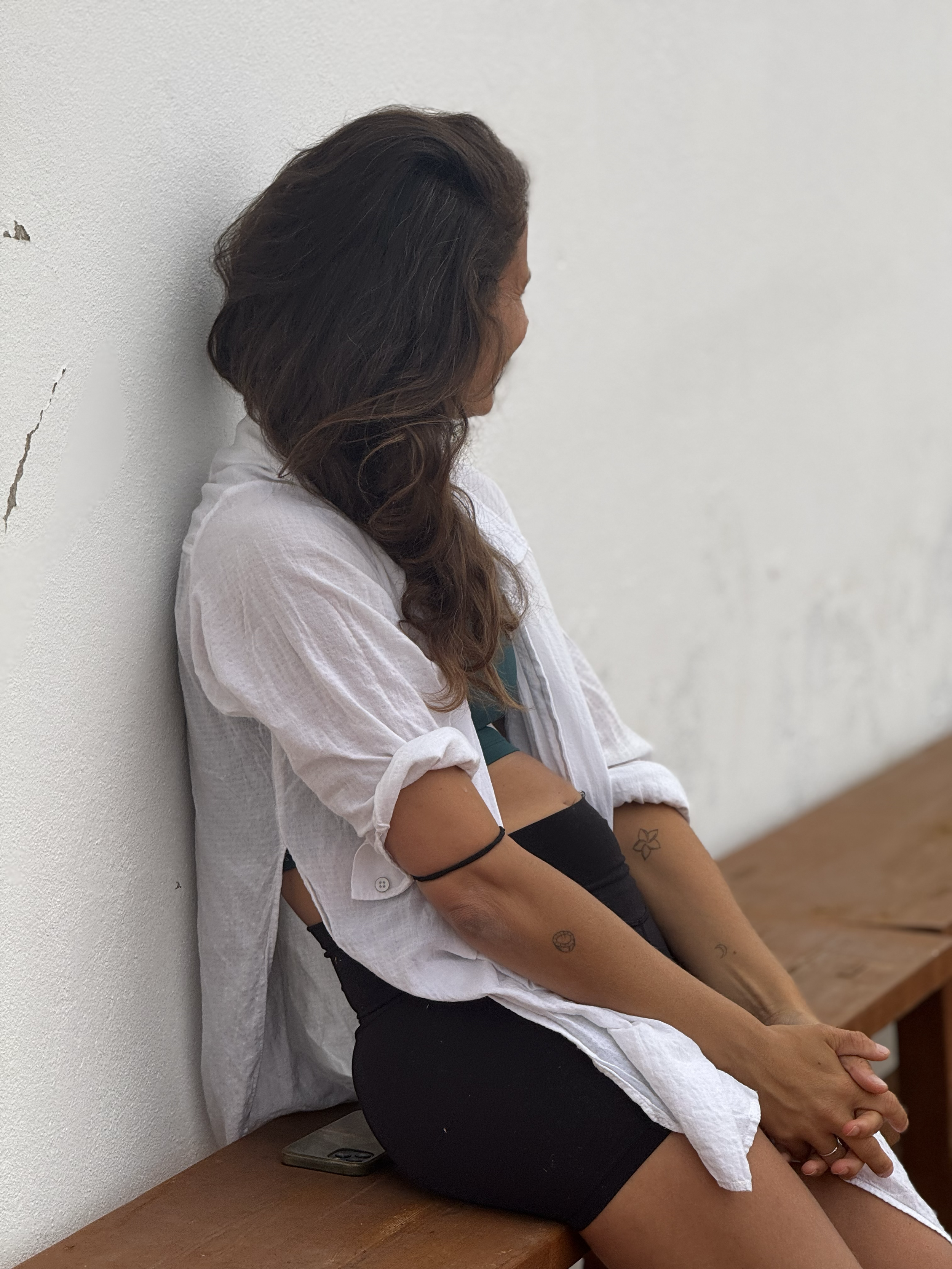 A woman with dark, wavy hair sitting on a wooden bench, wearing a white shirt with rolled-up sleeves, a green top, and black shorts. She has tattoos on her arms and is looking down with her hands clasped in her lap, next to a smartphone on the bench.