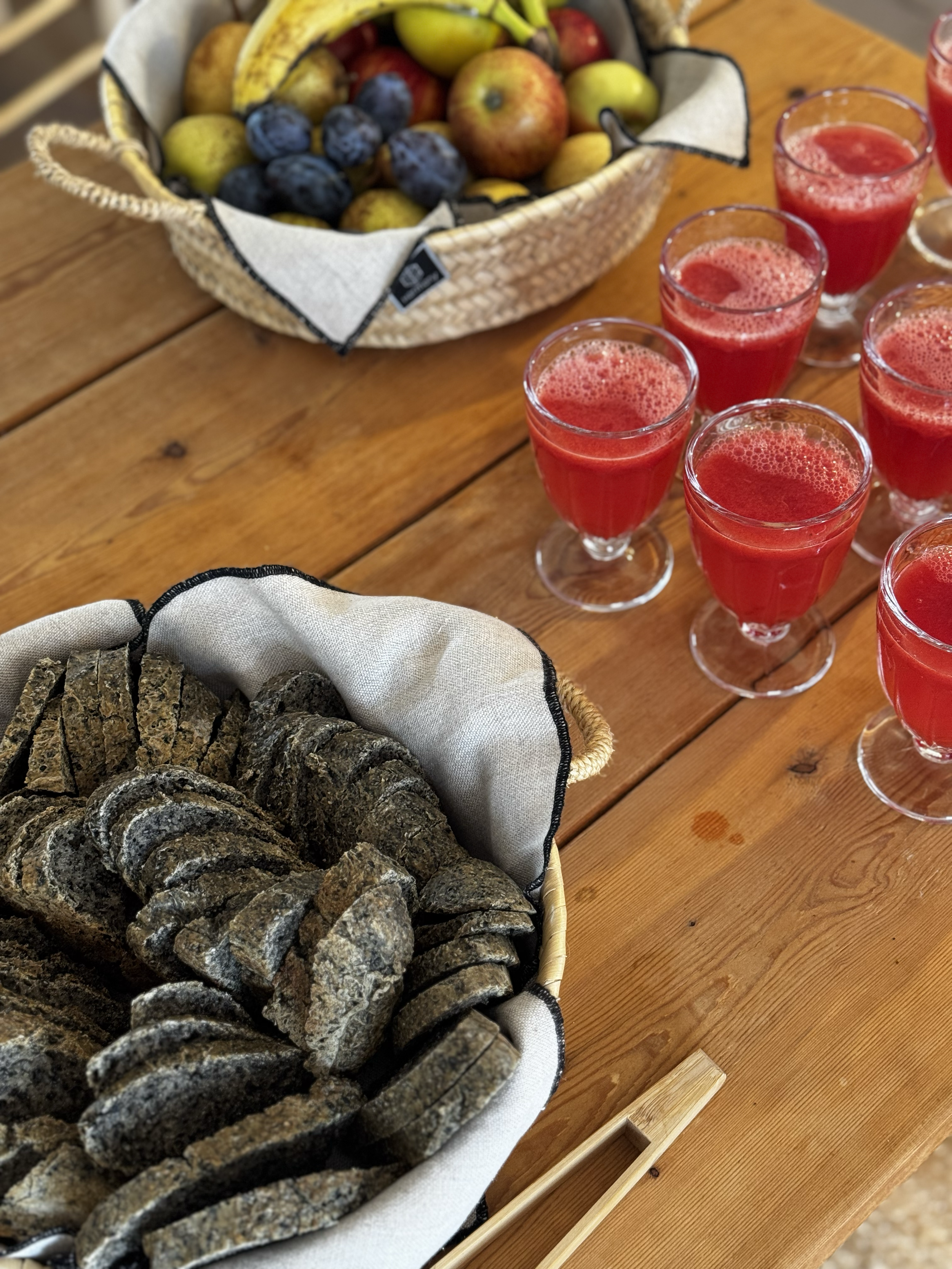 A wooden table with a basket of fresh fruit, a basket of sliced bread, and several glasses of red fruit juice at a women's only yoga retreat in Portugal