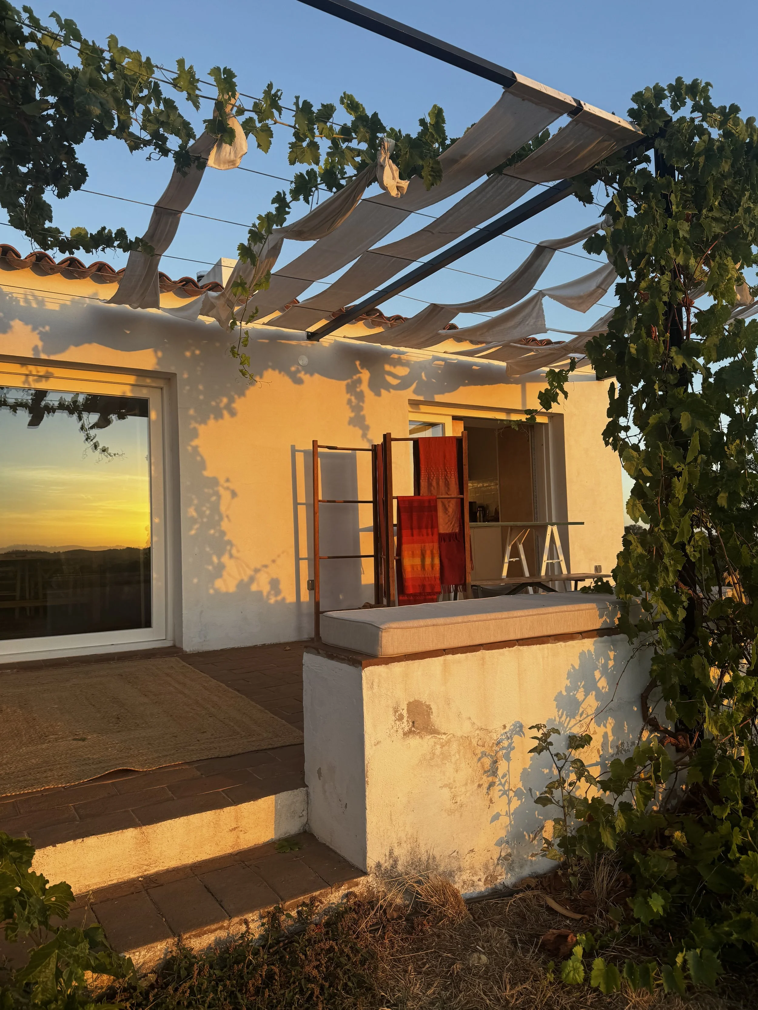 Sunset view of a the yoga retreat accommodation with white walls, a sliding glass door, a cushioned bench, and clothes hanging on a rack. Green vine plants grow around the terrace.
