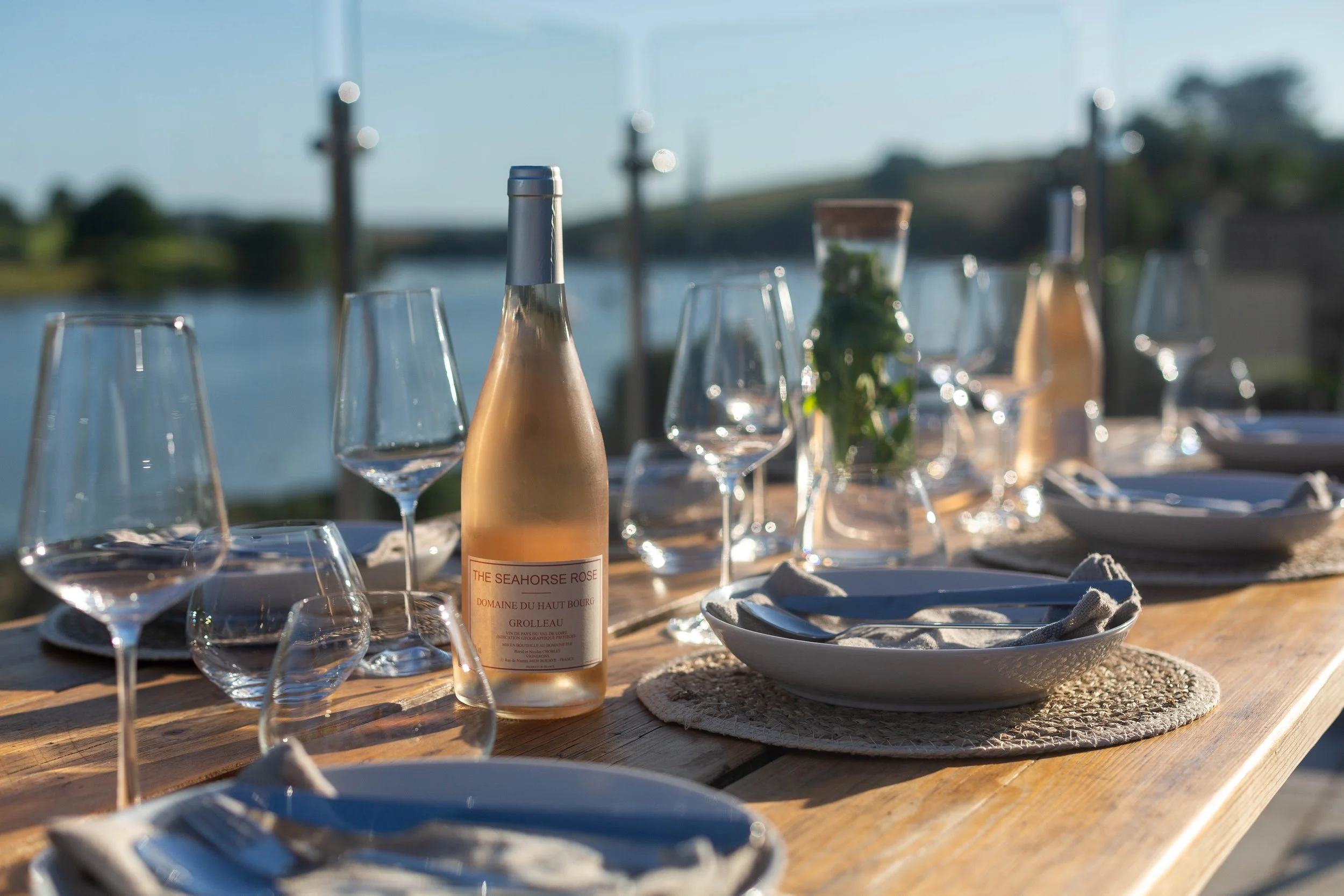 Outdoor dining table set with wine bottles, glasses, plates, and napkins near a river with a bridge in the background, during daytime for yoga retreat guests in Devon