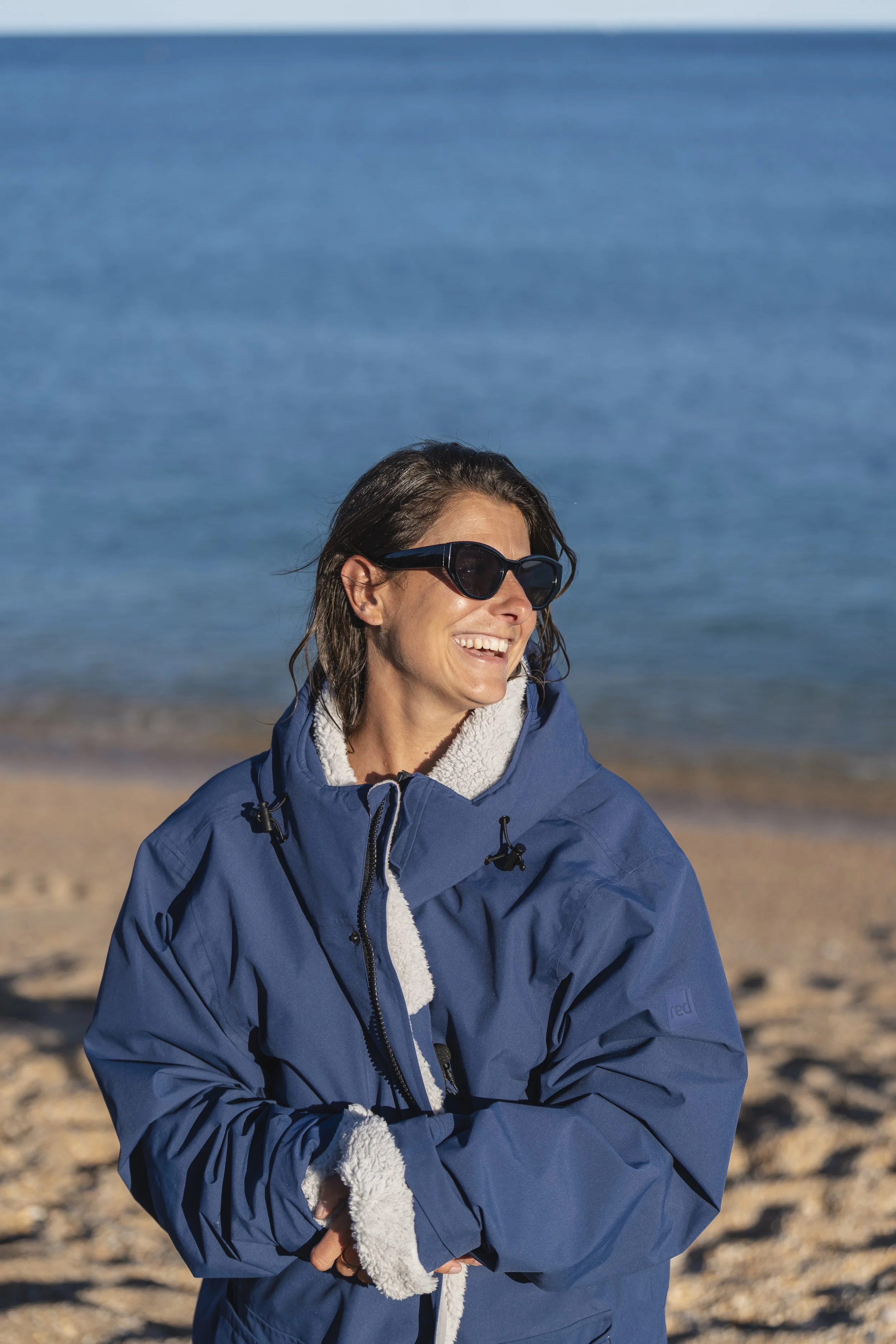 Woman in a blue jacket and sunglasses standing on a beach with the ocean in the background.