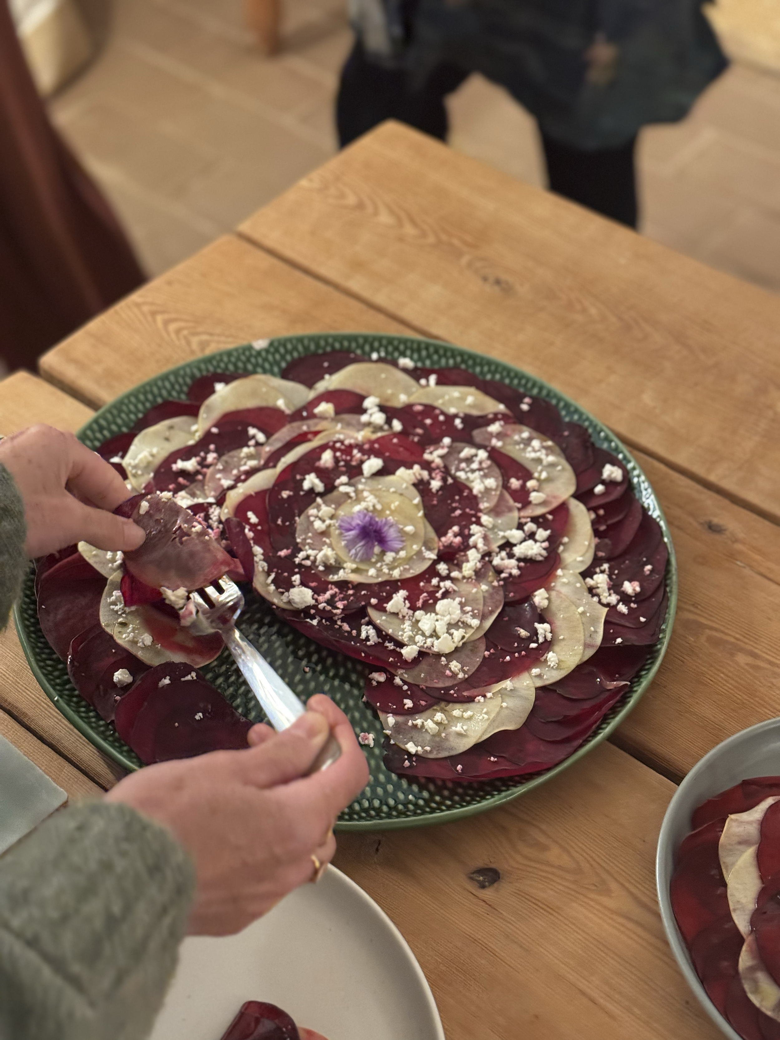 A person arranges a beet and goat cheese salad with edible flowers and crumbled cheese on a large green plate at a women's only yoga retreat in Portugal