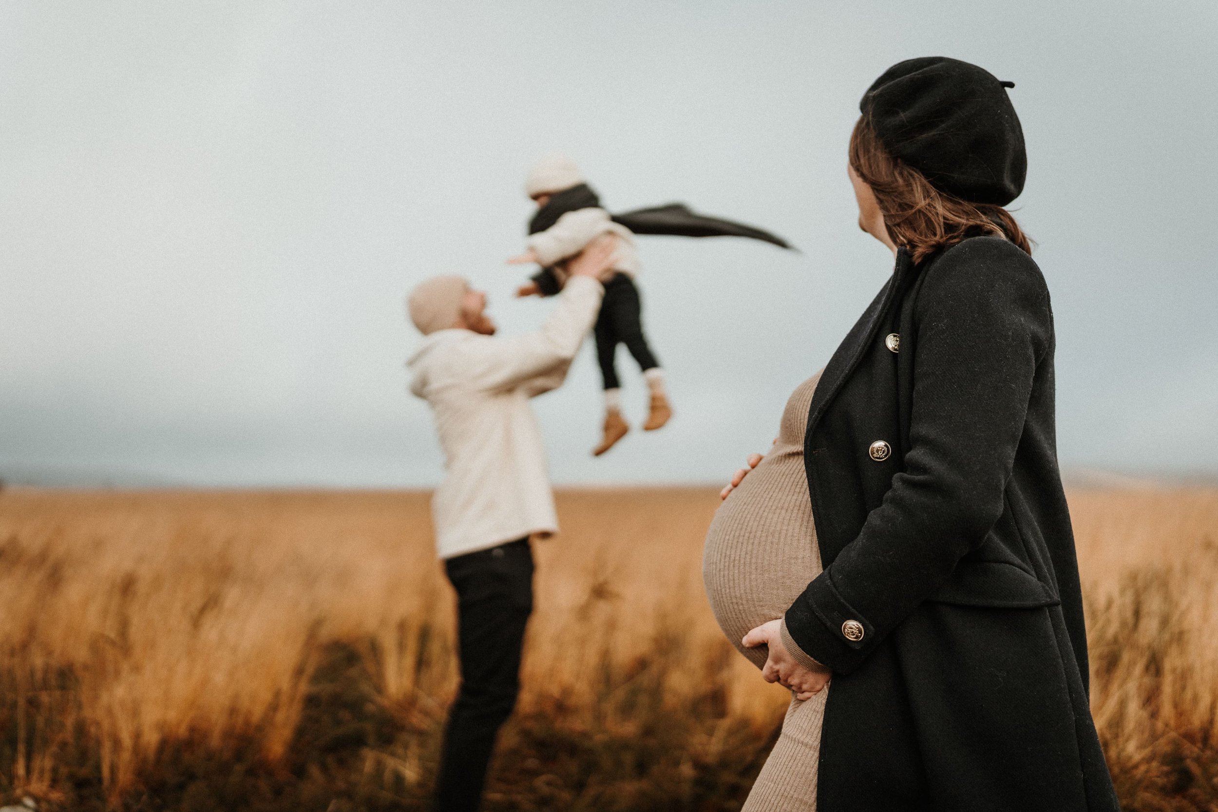 Séance famille lifestyle en extérieur, futur papa soulève son enfant pendant qu’une femme enceinte les regarde