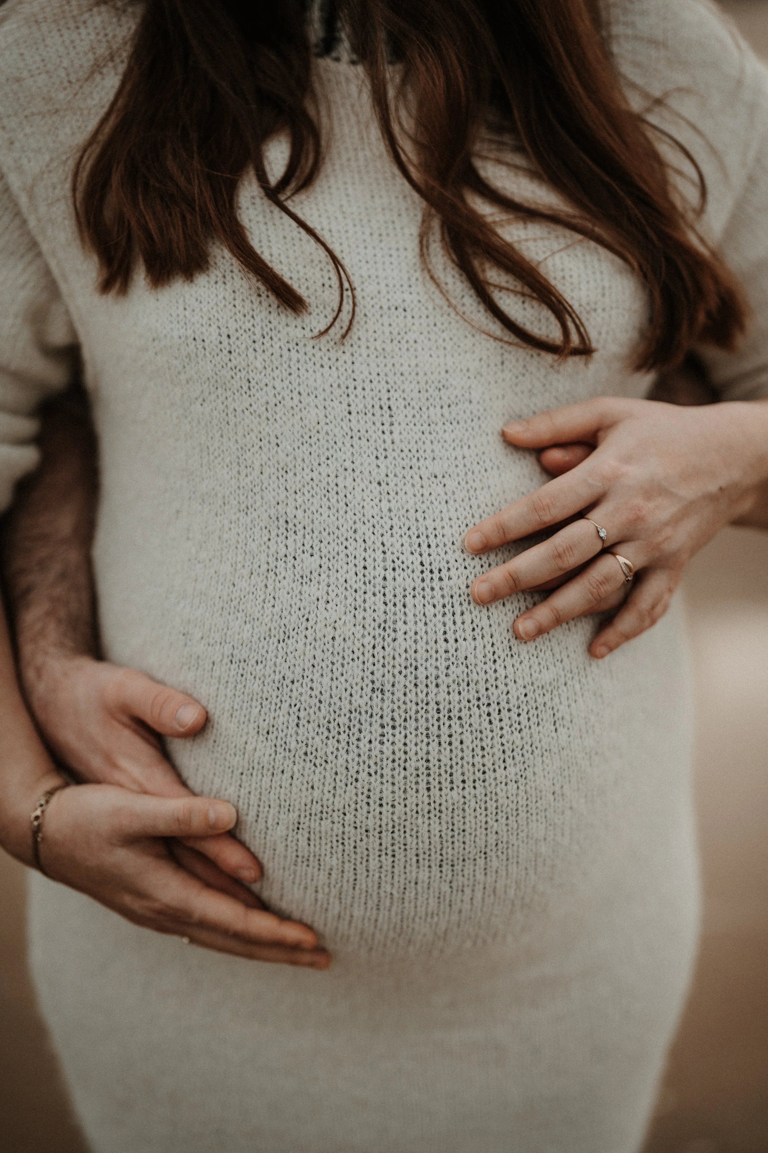 Ventre de femme enceinte entouré des mains des futurs parents lors d’une séance grossesse lifestyle