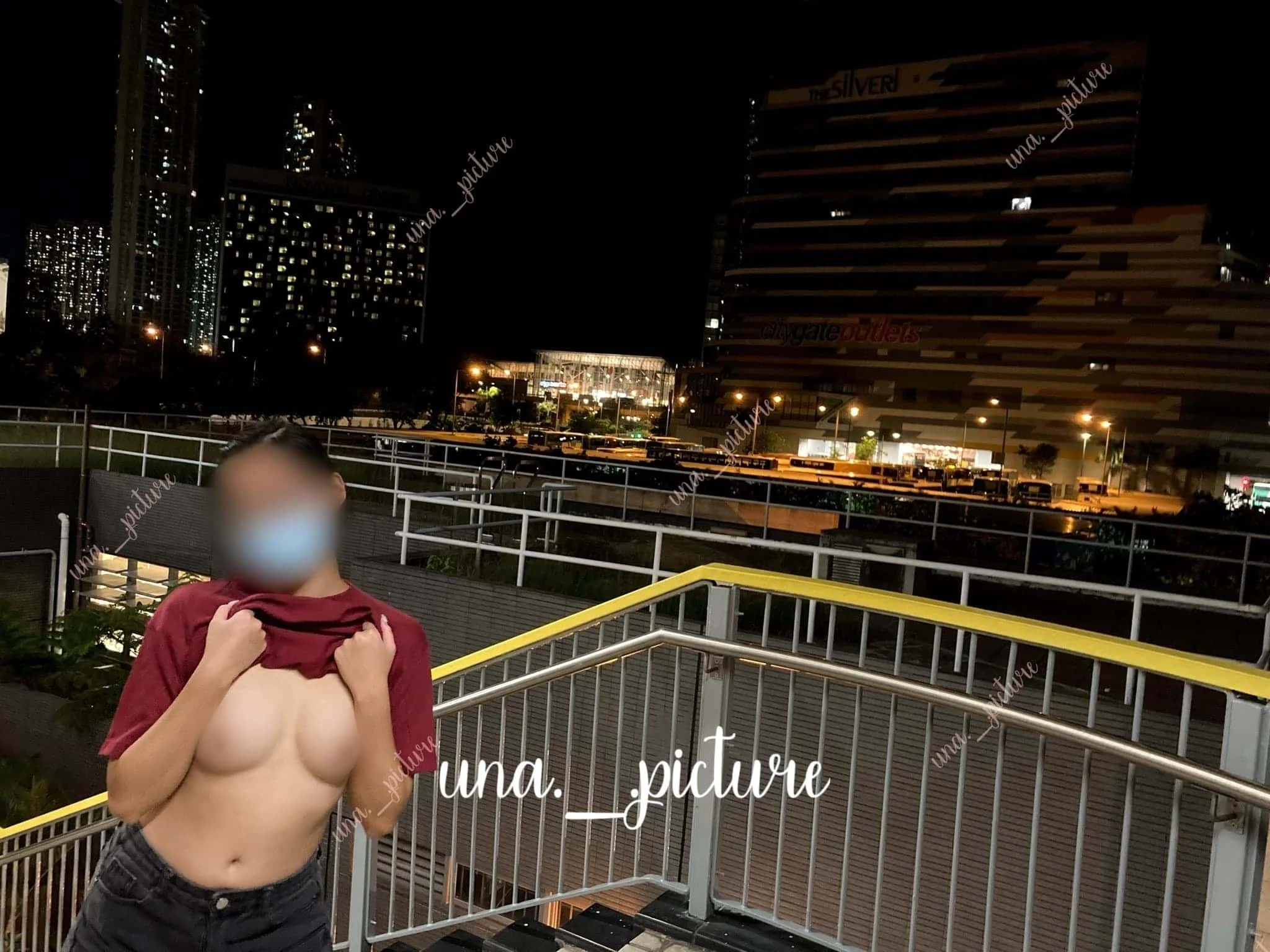 A person lifting their shirt to reveal their chest and wearing a face mask, standing on an outdoor platform at night with a cityscape, including tall buildings and lit windows, in the background.