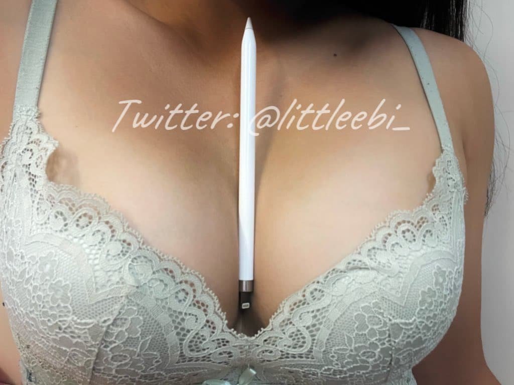 Close-up image of a woman's bare shoulders and cleavage wearing a lace dress with a white pen placed vertically between her breasts, and a watermark text of Twitter handle '@littl3ebi_' across the chest.