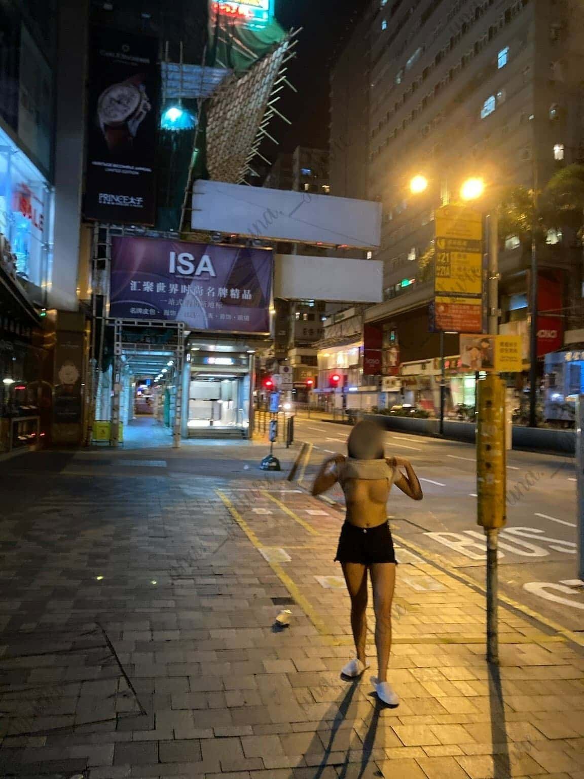 A woman with dark, curly hair walking on an empty city street at night, illuminated by streetlights and signs, with a purple advertisement banner and various buildings on either side.