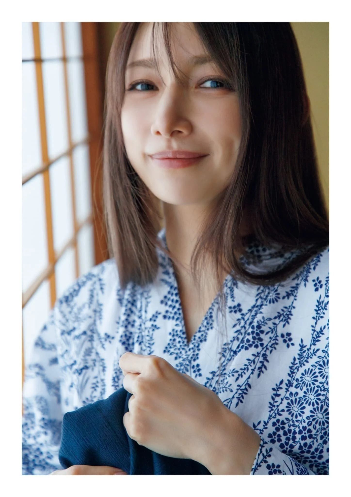 A young woman with shoulder-length brown hair, blue eyes, and fair skin, smiling softly, holding a dark jacket or sweater, standing near a window with wooden lattice framing.