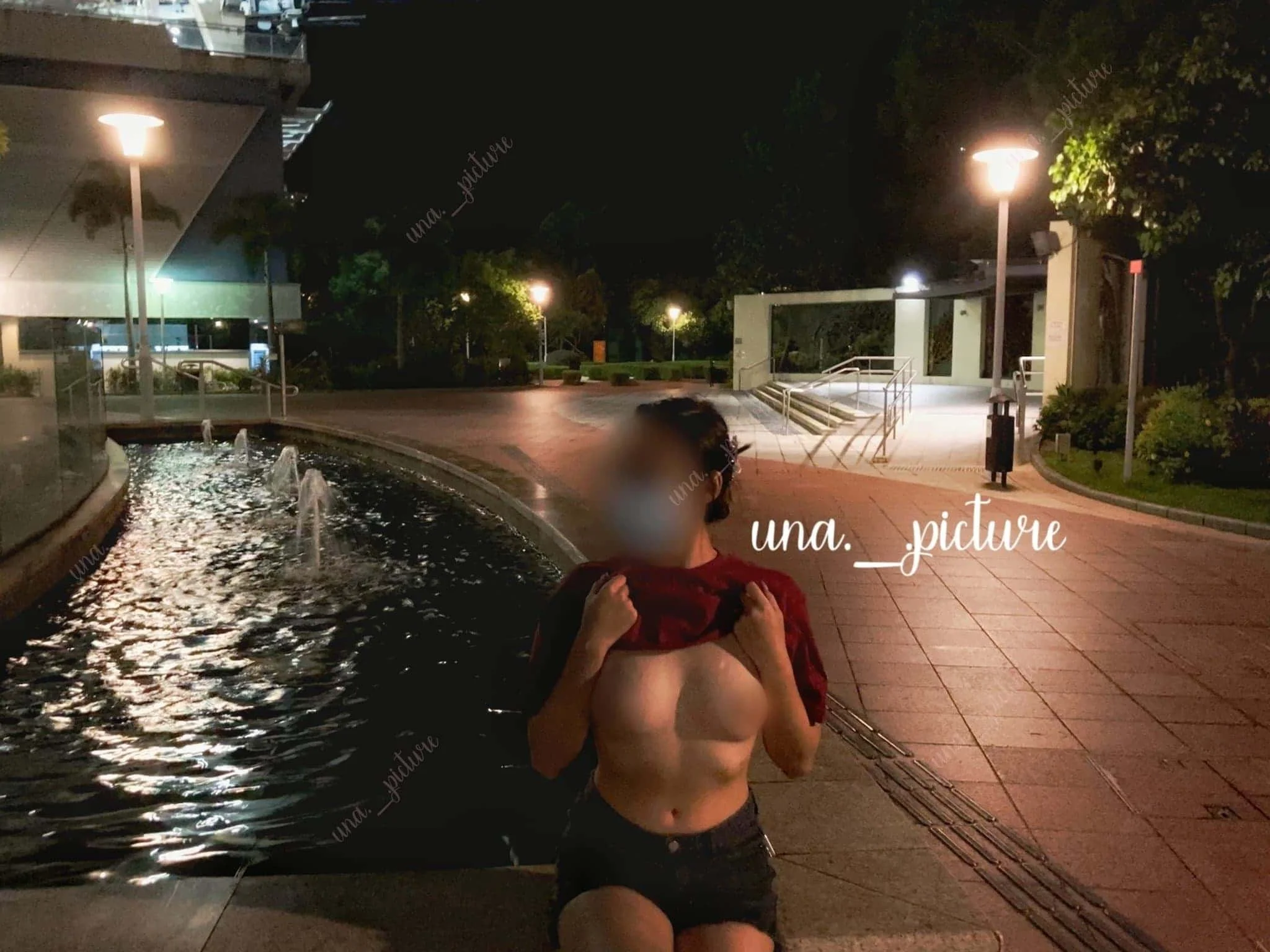 A person with blurred face, holding a red shirt off their shoulders, standing outdoors at night near a water fountain and tiled walkway, with street lamps illuminating the scene.