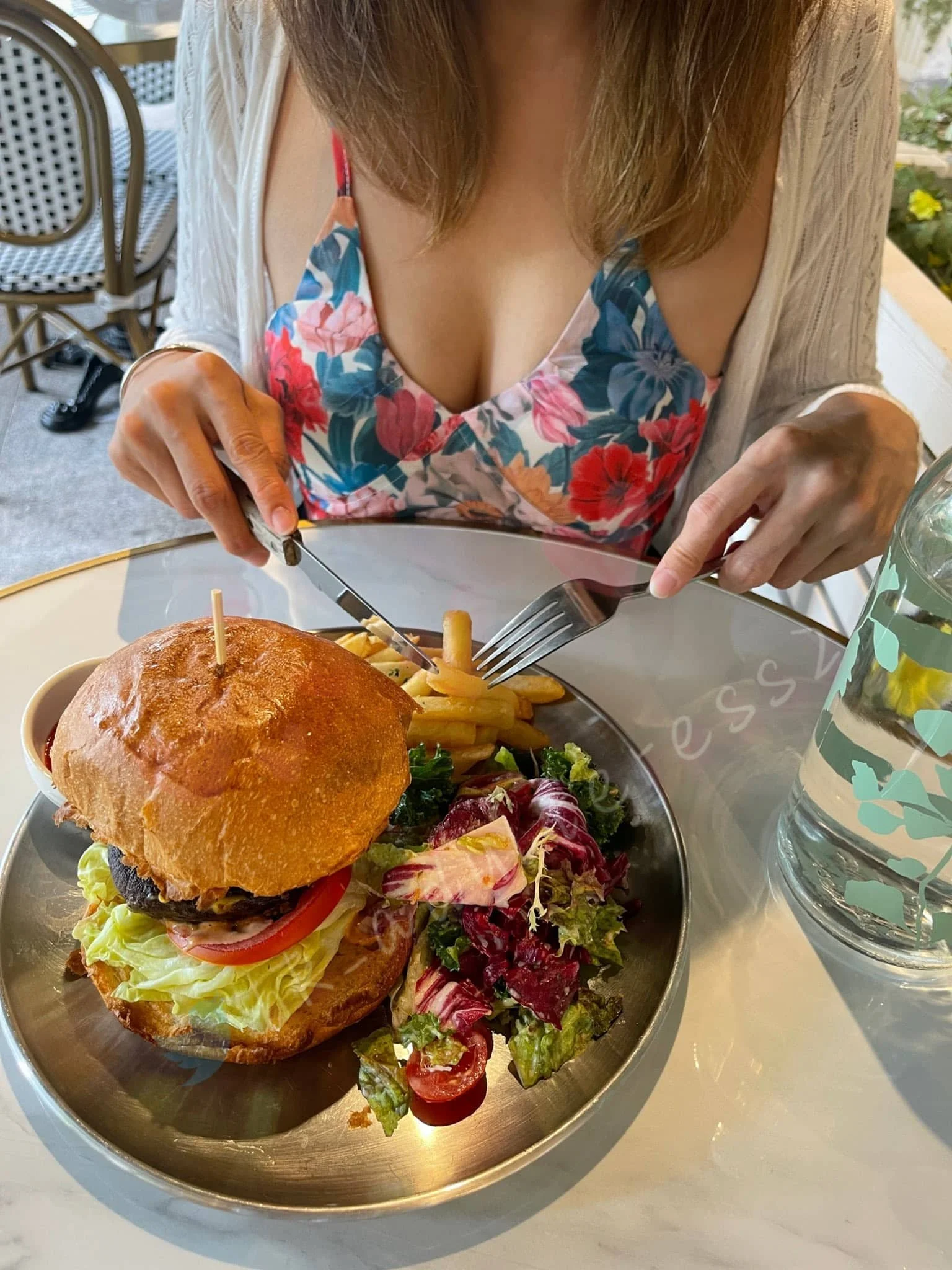 A woman cutting into a burger with French fries and salad on a stainless steel plate at a dining table.