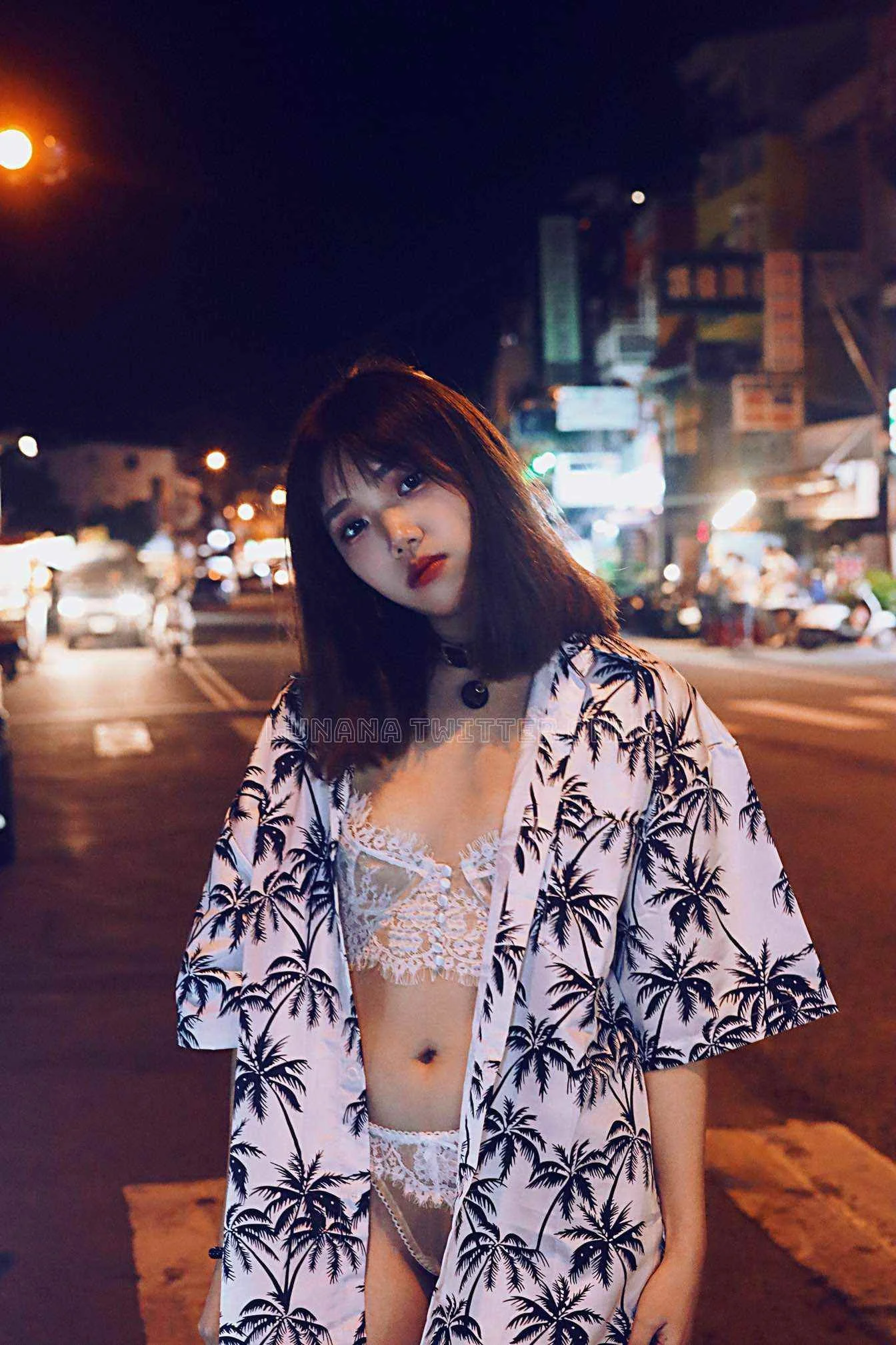 Young woman standing on a city street at night, wearing a white lace bralette and shorts, with a black choker, and a tropical print button-up shirt with palm trees, illuminated by city lights in the background.