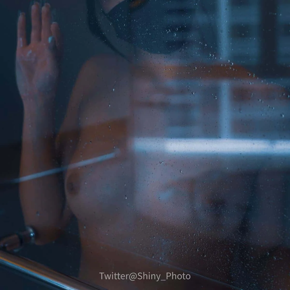 A person with blond hair pressing their face and hand against a wet glass surface, with droplets of water on the glass. The person's upper body is visible behind the glass.