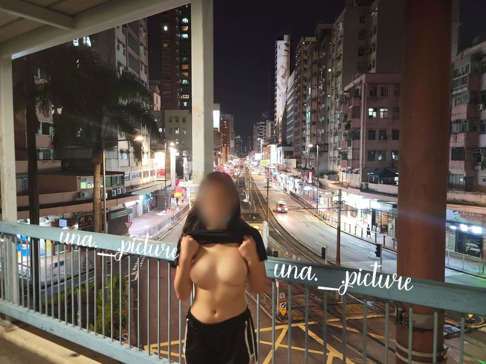 A person standing topless on a bridge at night, with a city street and tall buildings in the background.
