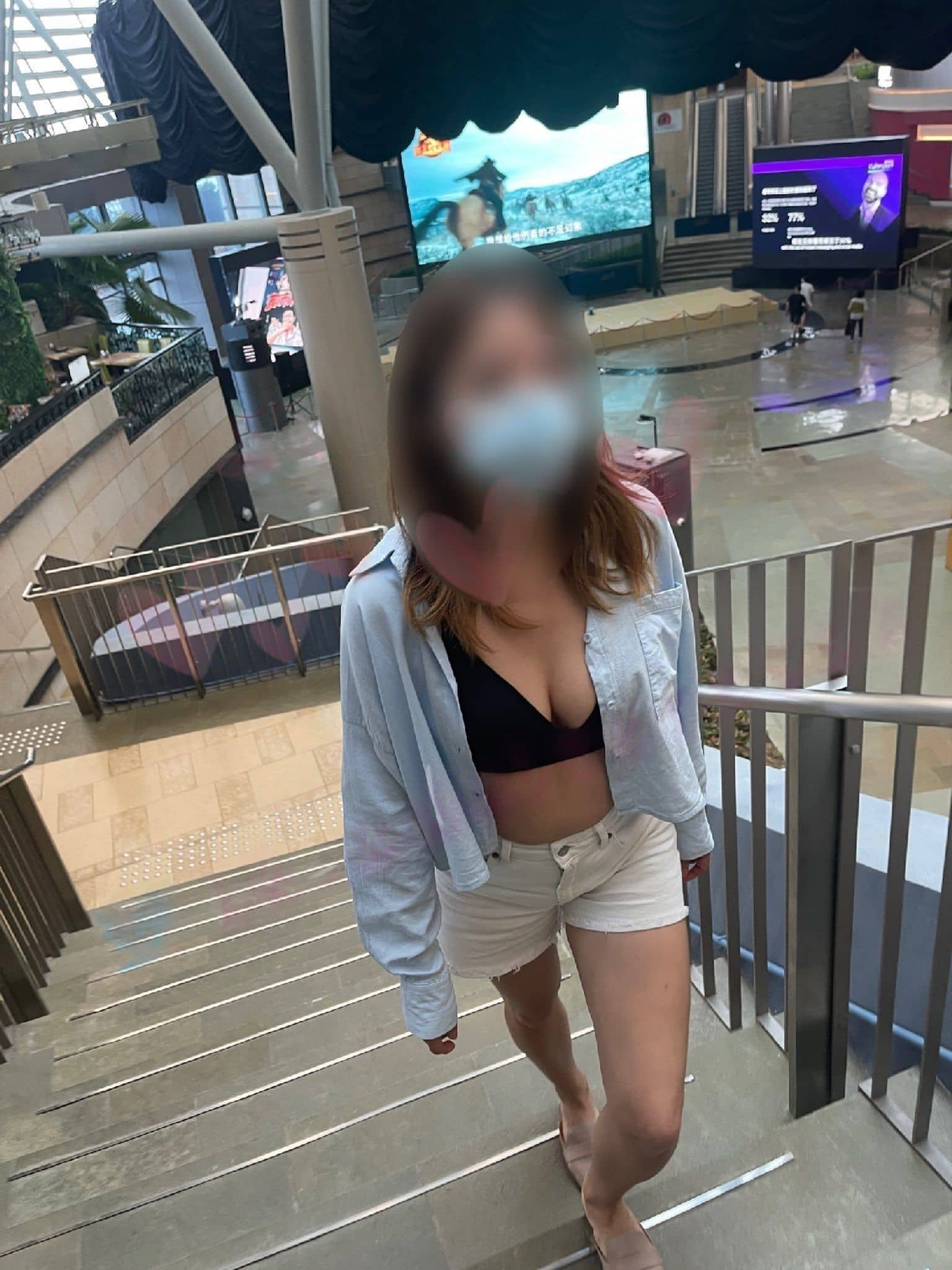 A woman wearing a mask, black top, an open light blue shirt, and white shorts, walking up a staircase in an indoor shopping mall or entertainment complex.