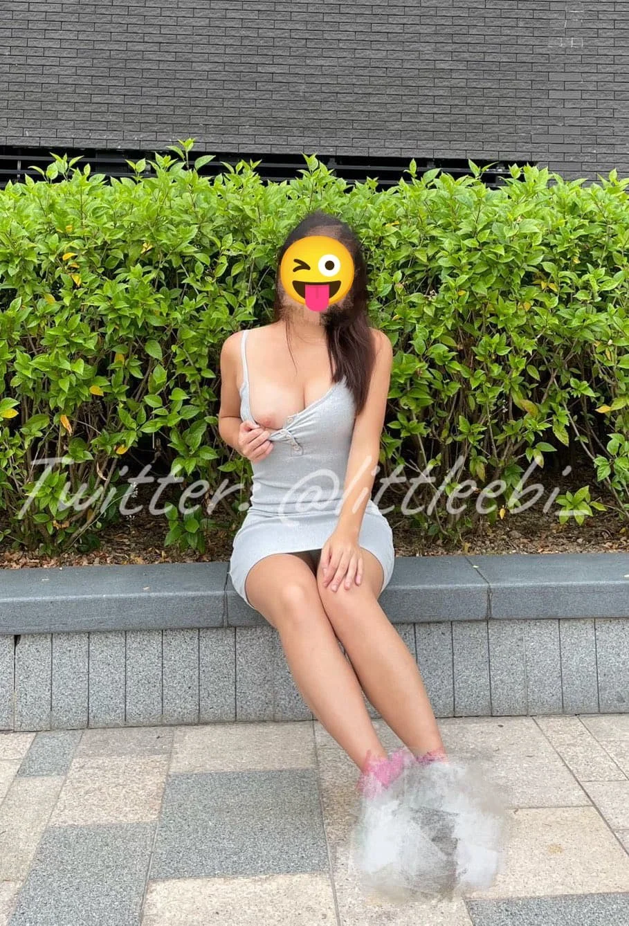 A woman with an emoji face covering sits on a bench with green bushes in the background and water splashing near her feet.