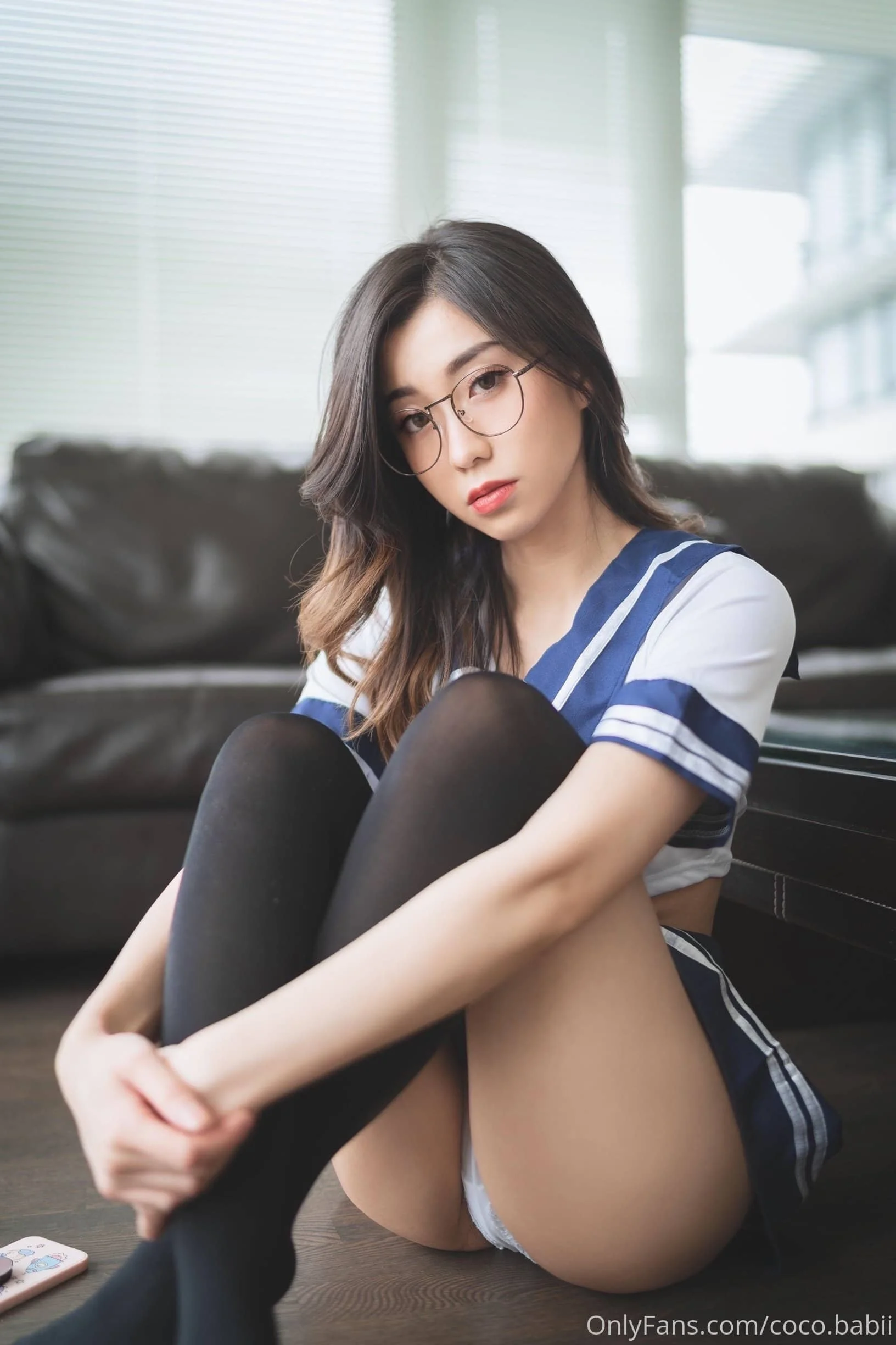 Person in a sailor-style outfit, wearing glasses, sitting indoors with crossed legs on the floor.