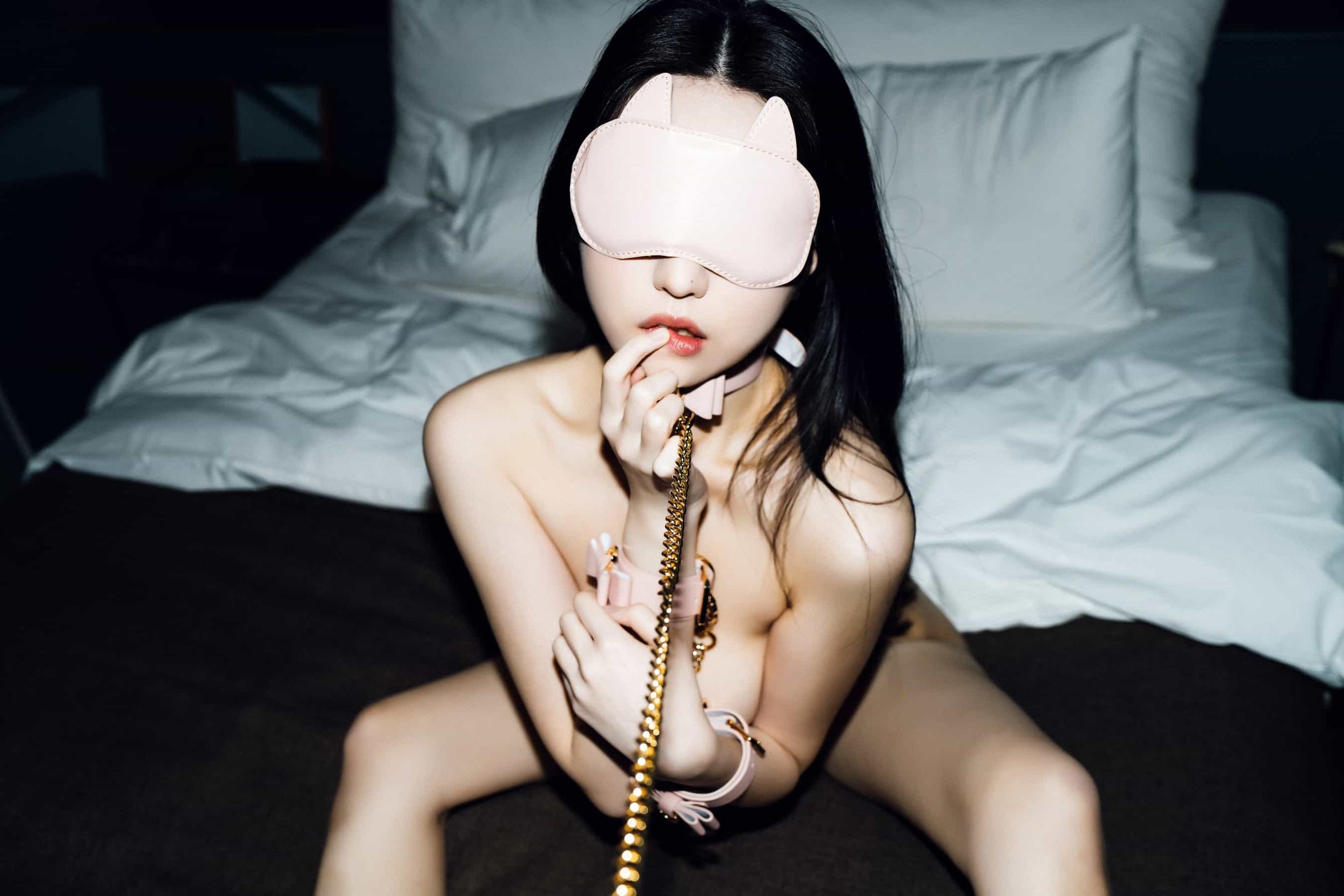 A woman with long dark hair is sitting on a bed, wearing a pink sleep mask with animal ears, holding a pink telephone receiver with a gold cord, and biting her finger, with white bedding in the background.