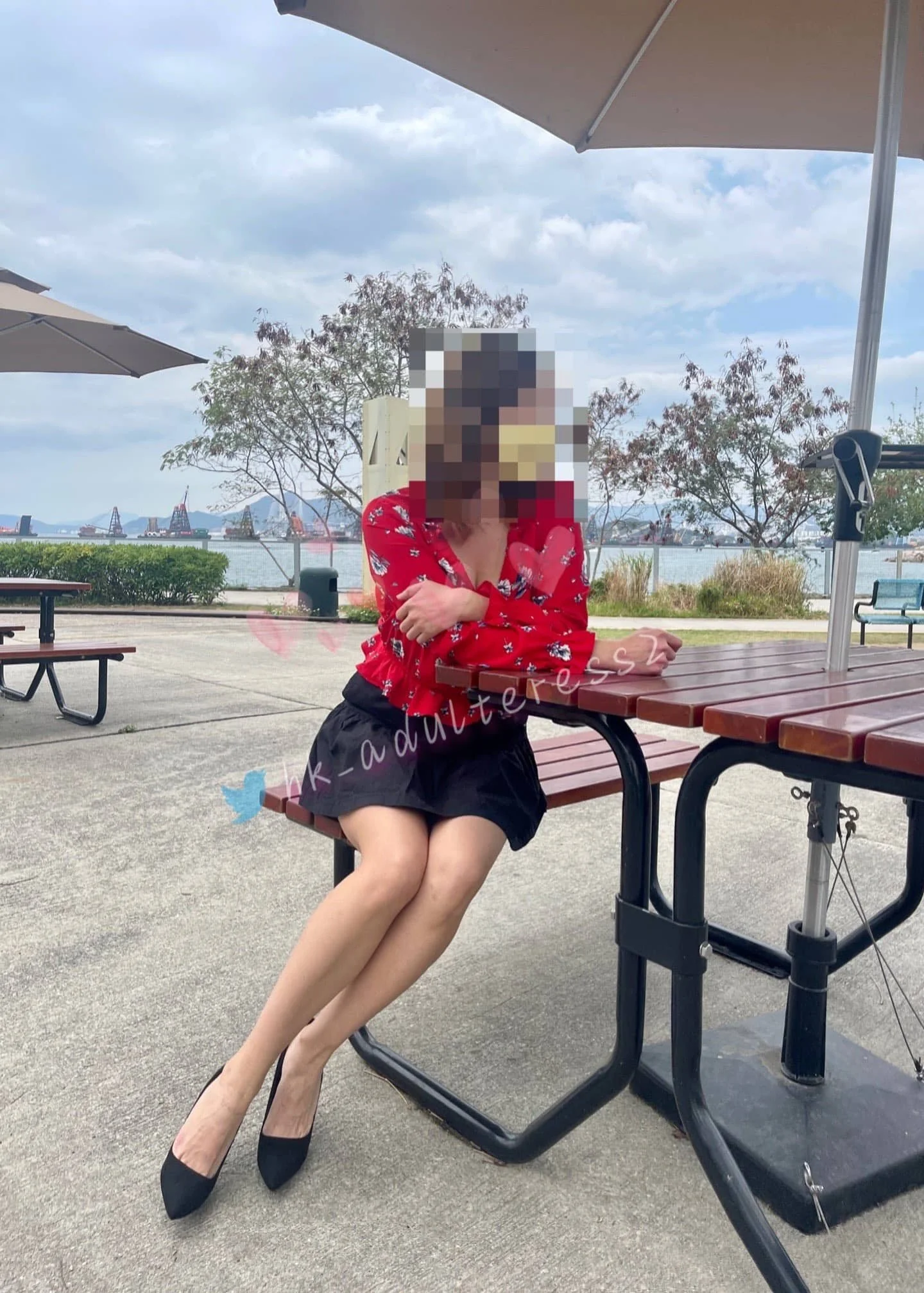 A person with a pixellated face sitting on a bench under a large umbrella at an outdoor location with picnic tables, trees, and water in the background.