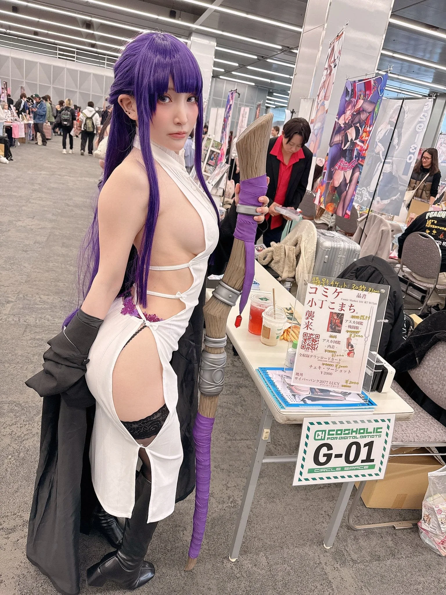 A woman in a cosplay costume with long purple hair, dressed in a revealing white outfit with thigh-high black stockings, holding a large wooden spear with purple accents at a convention booth with various posters and displays, and other attendees in 