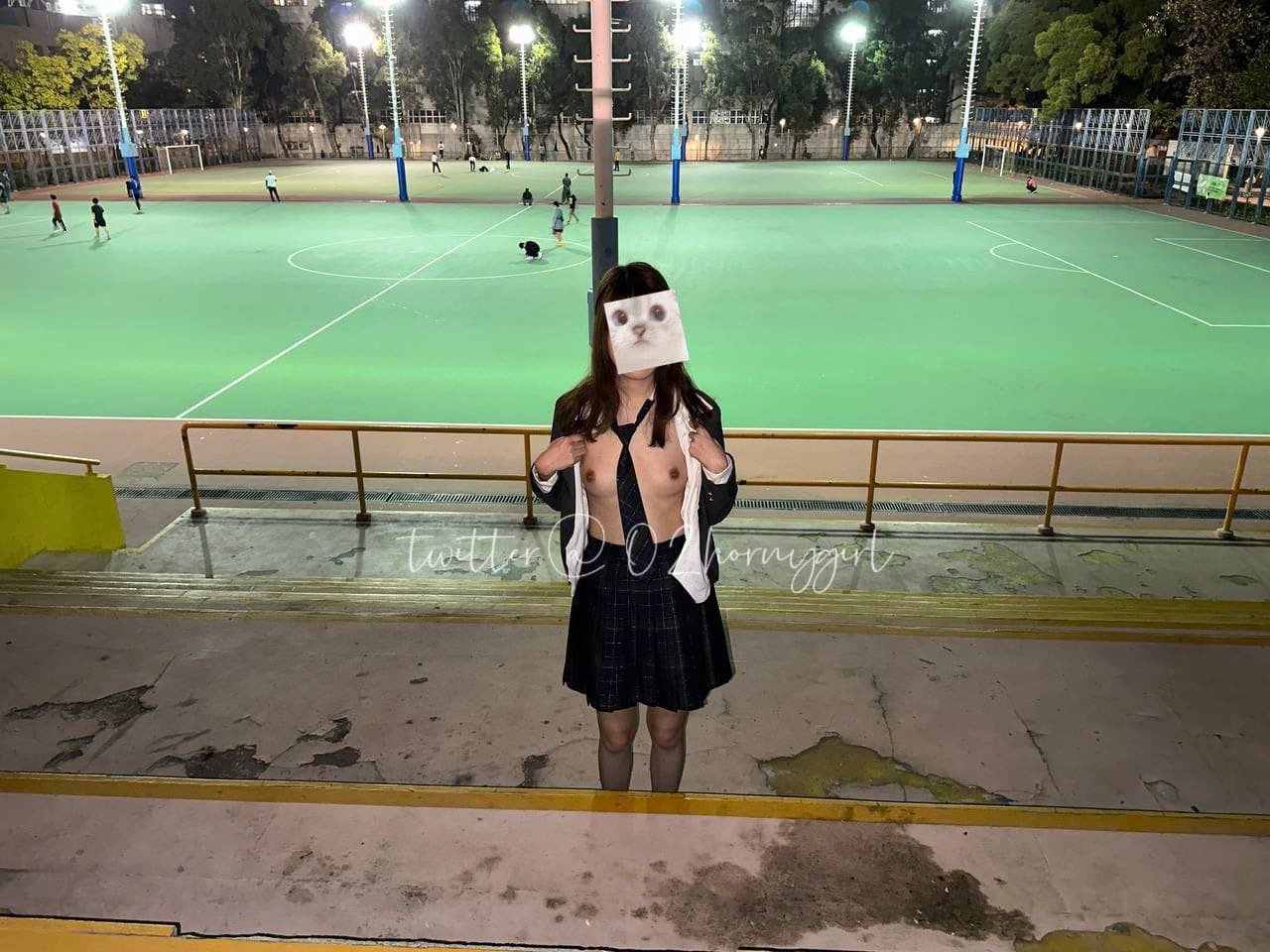 A person wearing a school uniform and a paper mask with a cat face is standing in front of an empty outdoor sports field at night, with bright lights illuminating the scene.