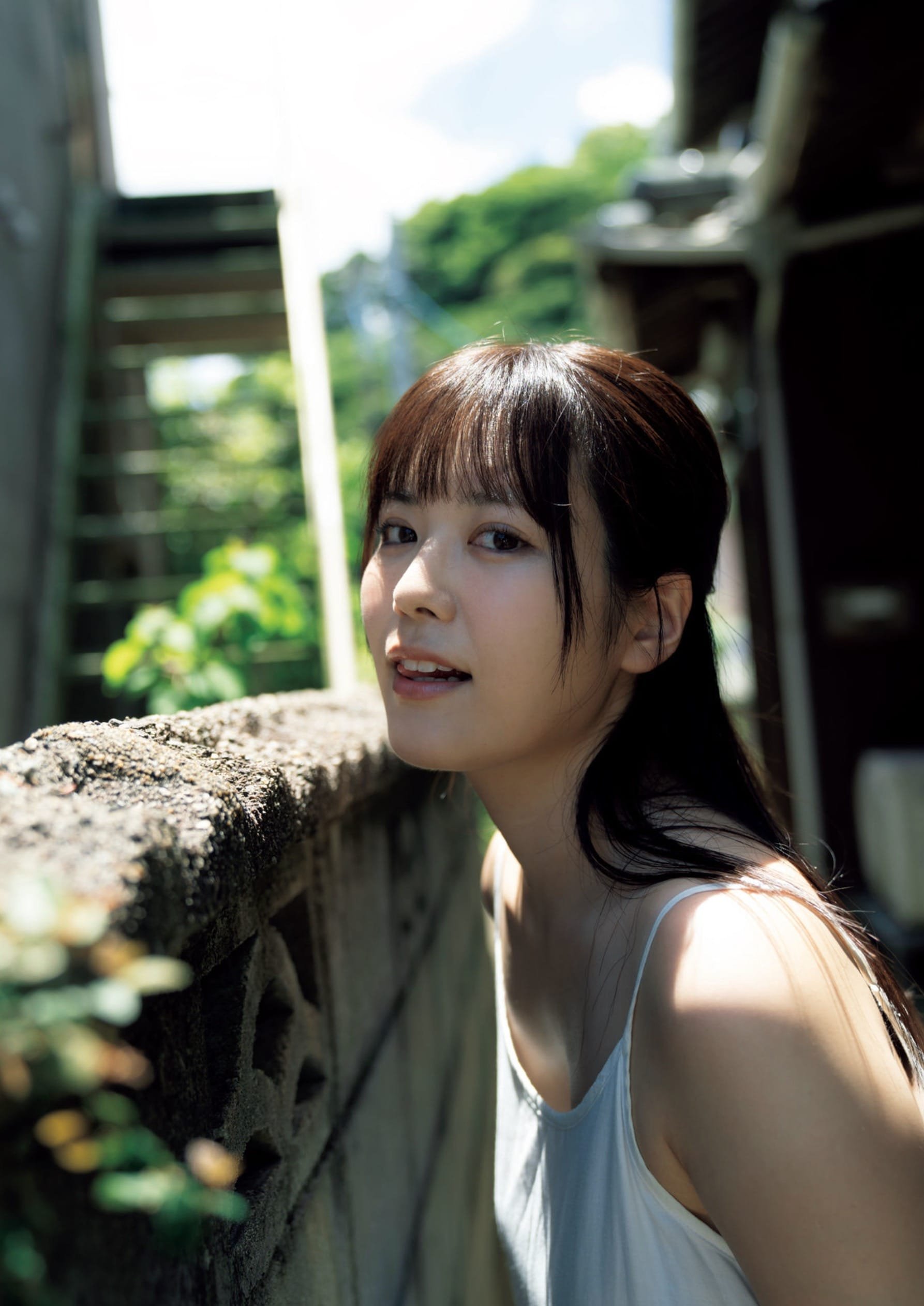A young woman with dark hair and a light top leans against a brick wall outdoors with sunlight and greenery in the background.