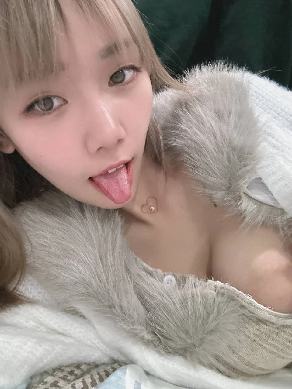 A young woman with light skin and light brown hair sticking out her tongue, wearing a fluffy white garment and a heart-shaped necklace, taking a selfie.