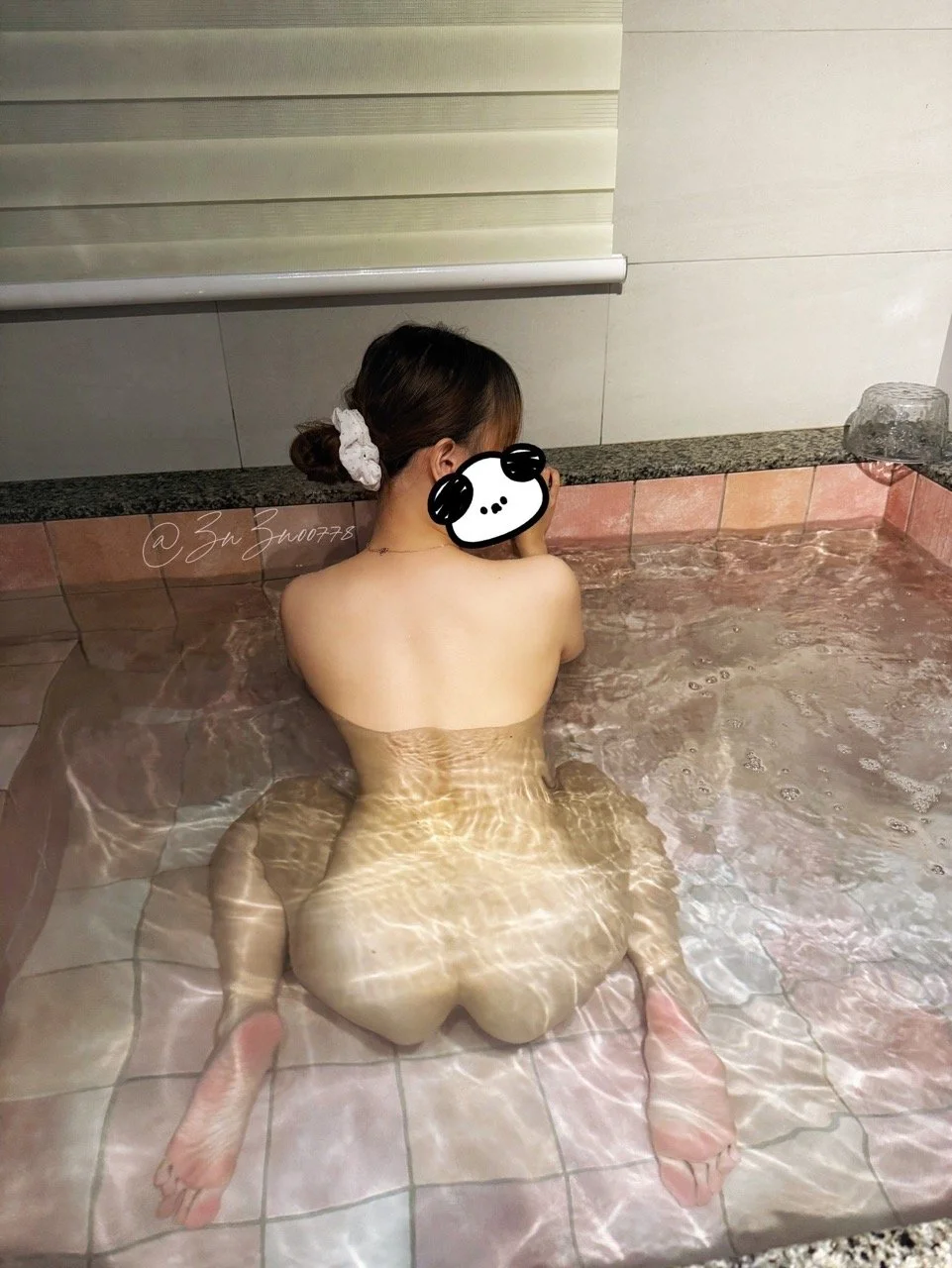 A woman kneeling in a bathtub with clear water, her back facing the camera, with her hair tied up with a white scrunchie. Her face is covered with a cartoon sticker. The bathtub has a tiled edge and a glass container on the right side.