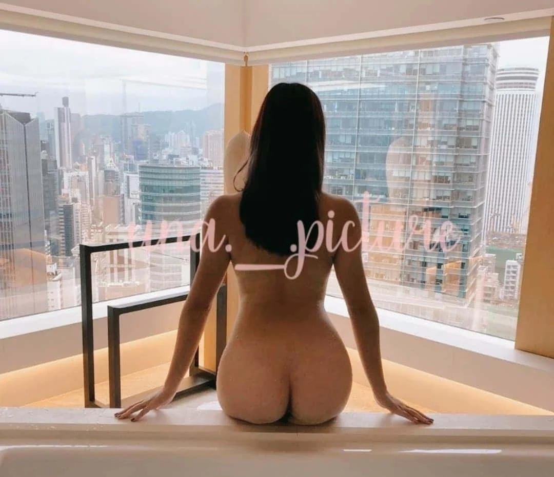 A woman with long dark hair sitting on the edge of a bathtub, facing large city skyscraper windows, with city buildings and mountains in the background.