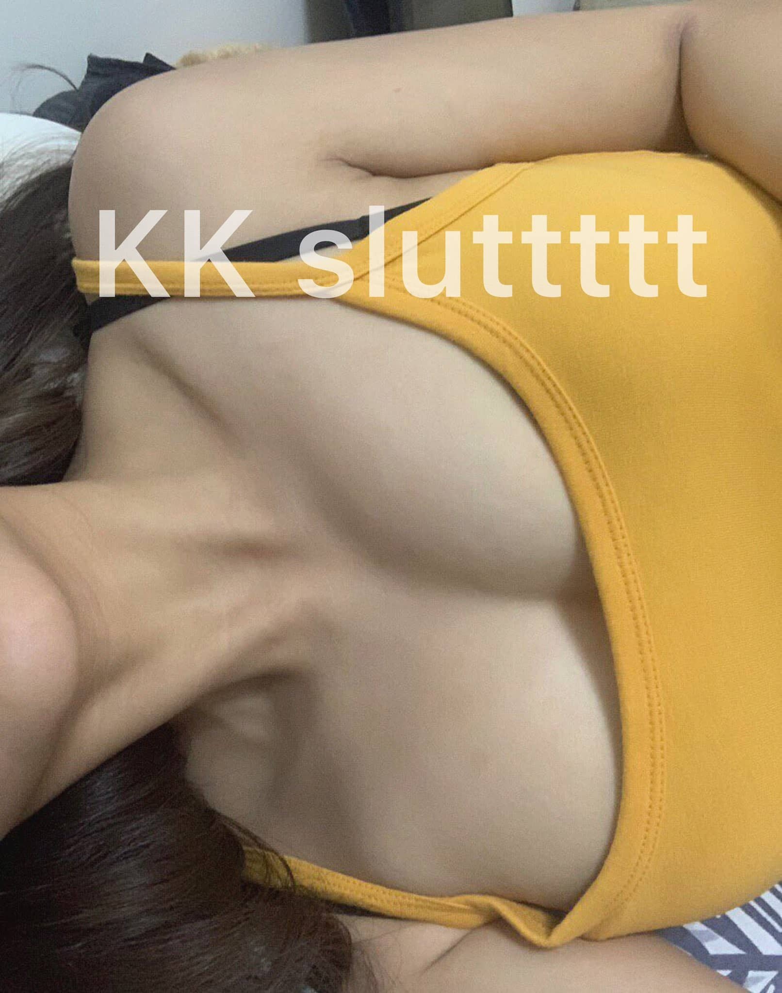 Close-up of a woman lying on her side on a bed, wearing a yellow tank top and black underwear, with text overlaid that reads 'KK sluttttt'.