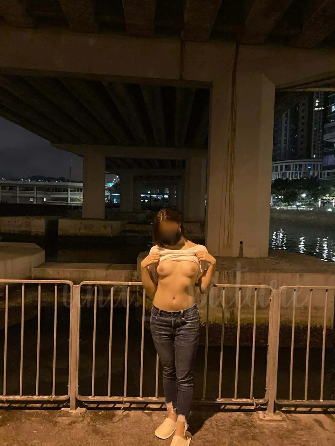 A woman standing outdoors at night, lifting her shirt to reveal her topless upper body, with a cityscape and water in the background.