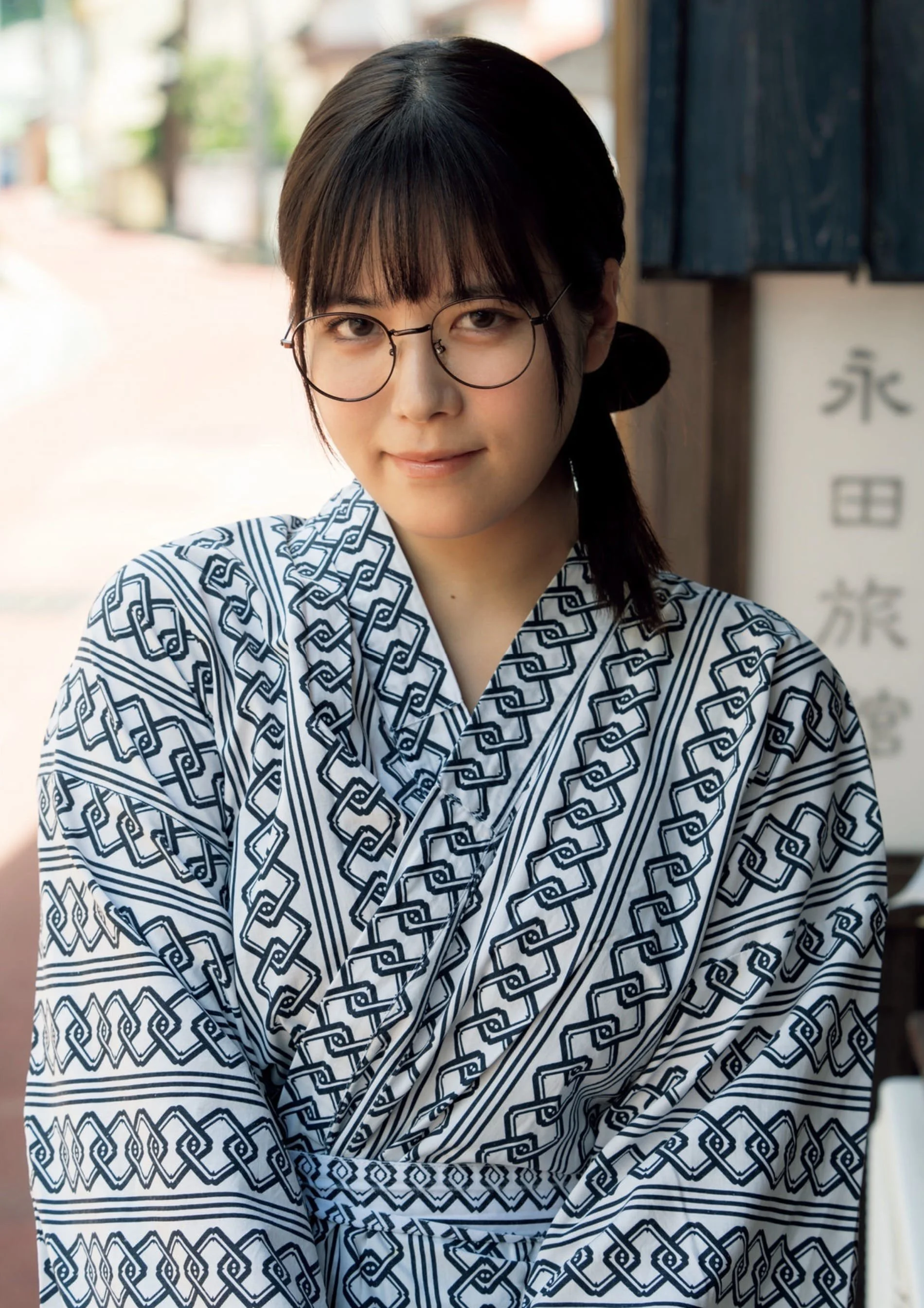 A young woman wearing glasses and a traditional Japanese yukata with a blue geometric pattern, standing outdoors with a slight smile.