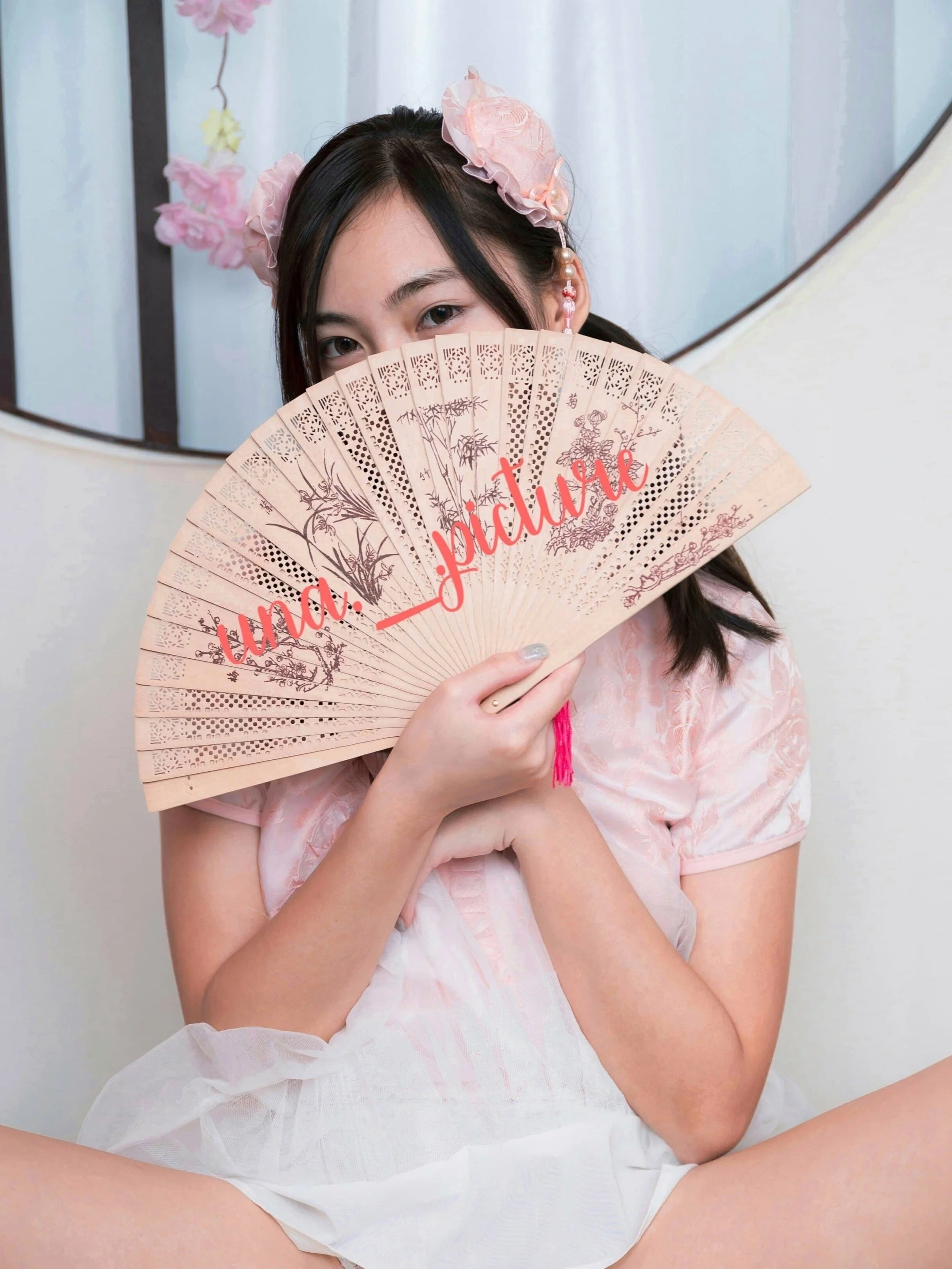 Person holding a decorative fan covering face, wearing a floral headband, white and pink outfit. Background with a mirror and hanging flowers.