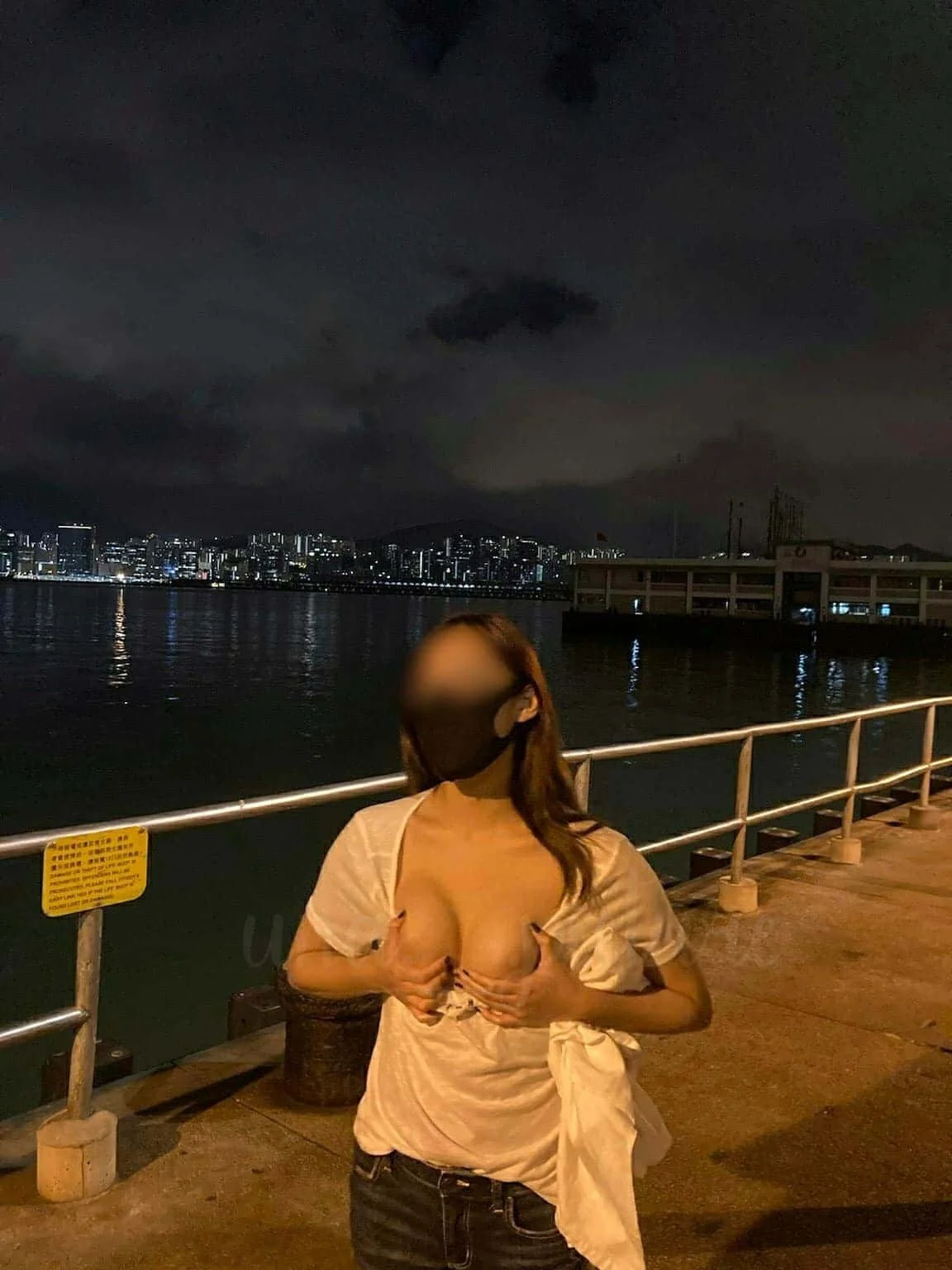 A woman standing on a waterfront at night, partially lifting her shirt and covering her chest with her hands. City skyline with lit buildings and dark clouds is in the background.