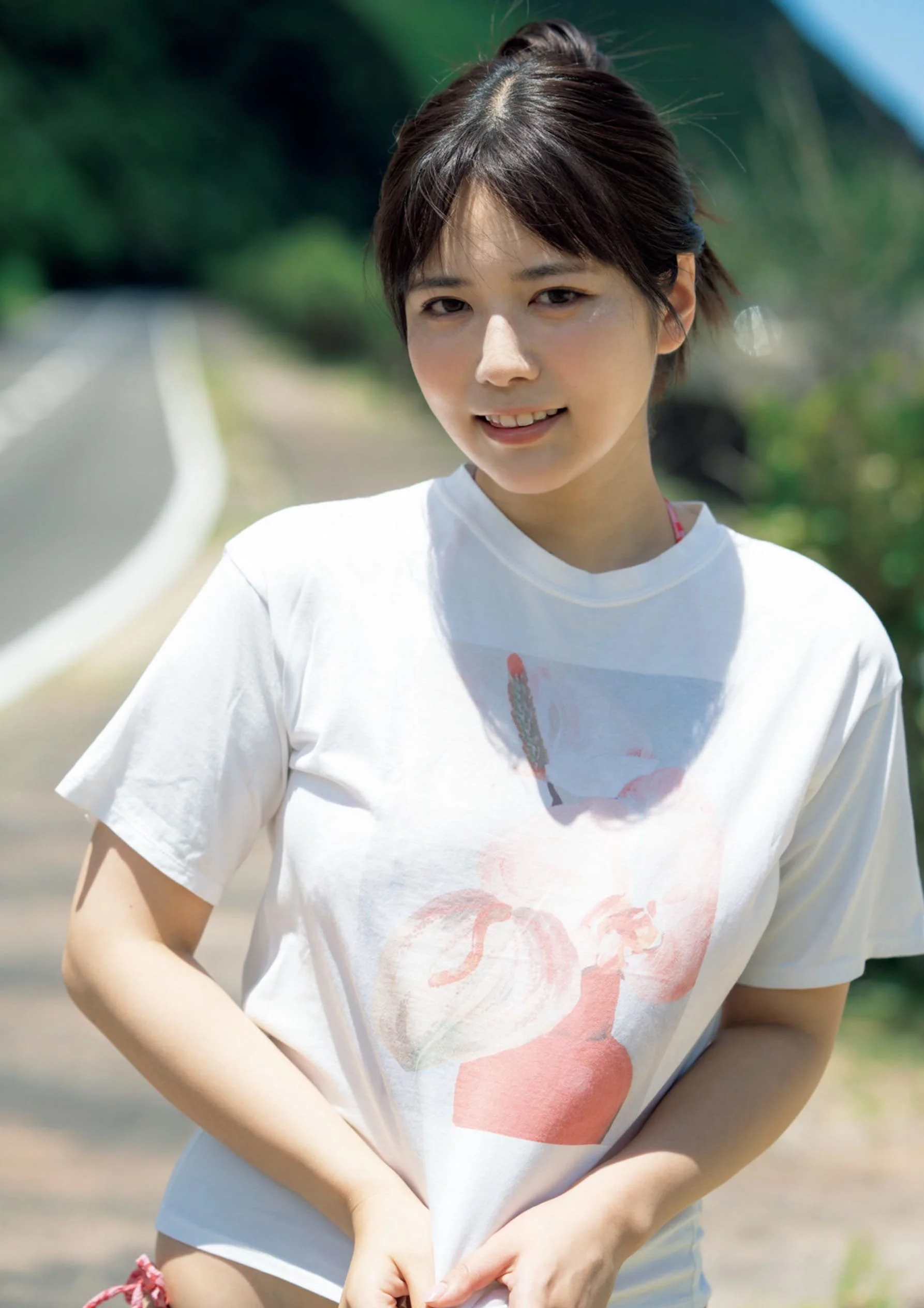 A young woman with short dark hair wearing a white T-shirt with a pink floral design, standing outdoors on a sunny day near a road with green trees in the background.