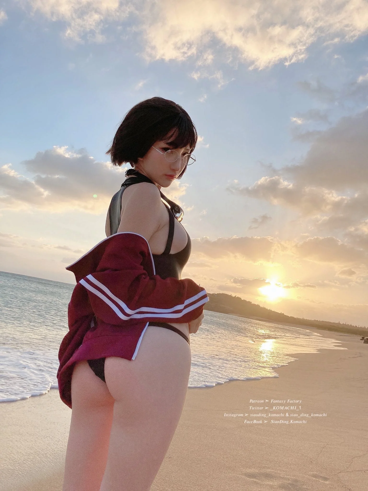 A woman with short dark hair and glasses standing on a beach at sunset, wearing a black bikini and a red jacket draped over her shoulders.