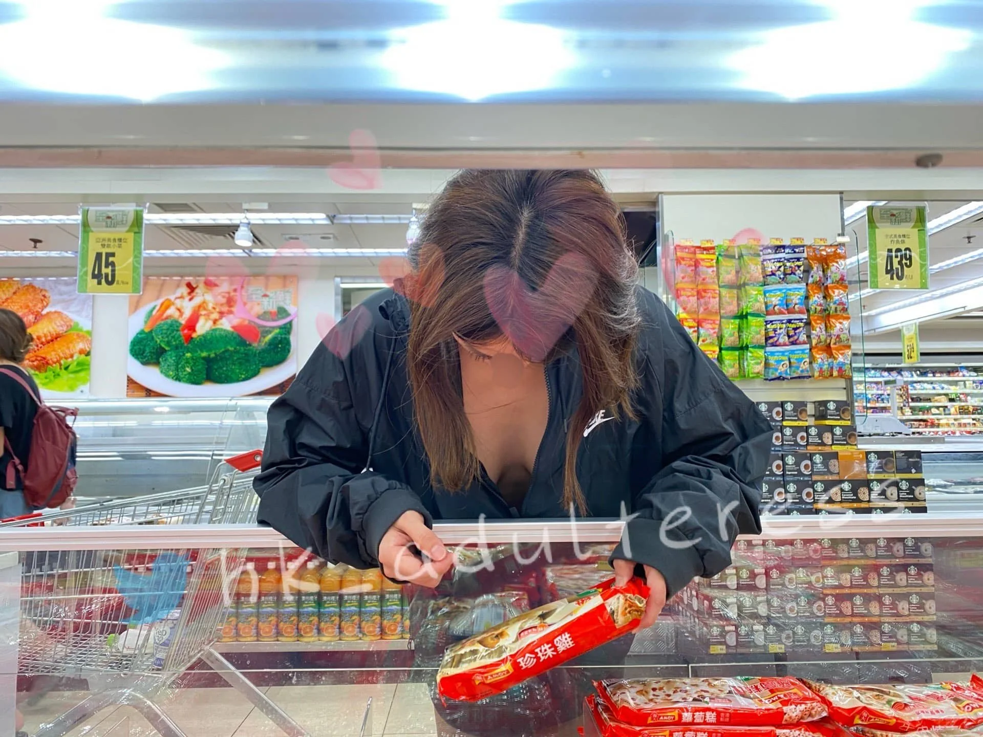 A woman leaning over a grocery store freezer, examining a package of food, with shelves of products and shopping cart in the background.