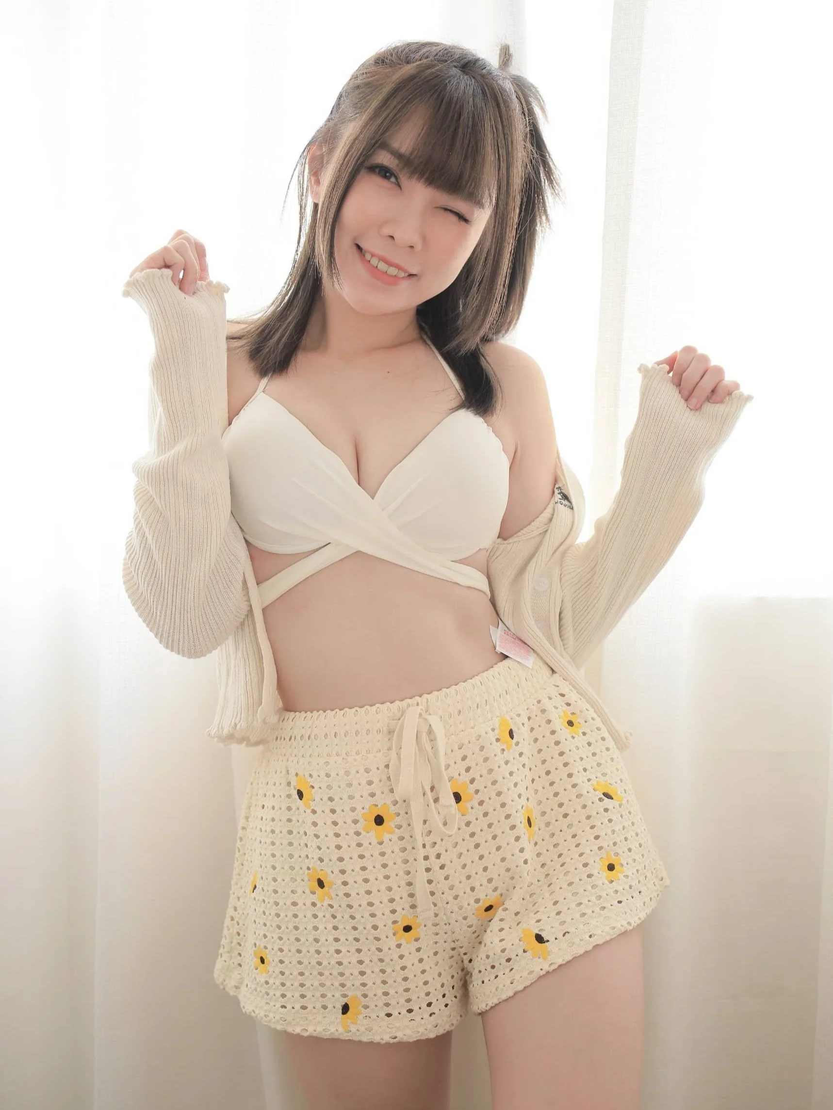 A person wearing a white bikini top, a white crochet cardigan, and cream crochet shorts with yellow flowers, standing in front of a light background, smiling and posing playfully.