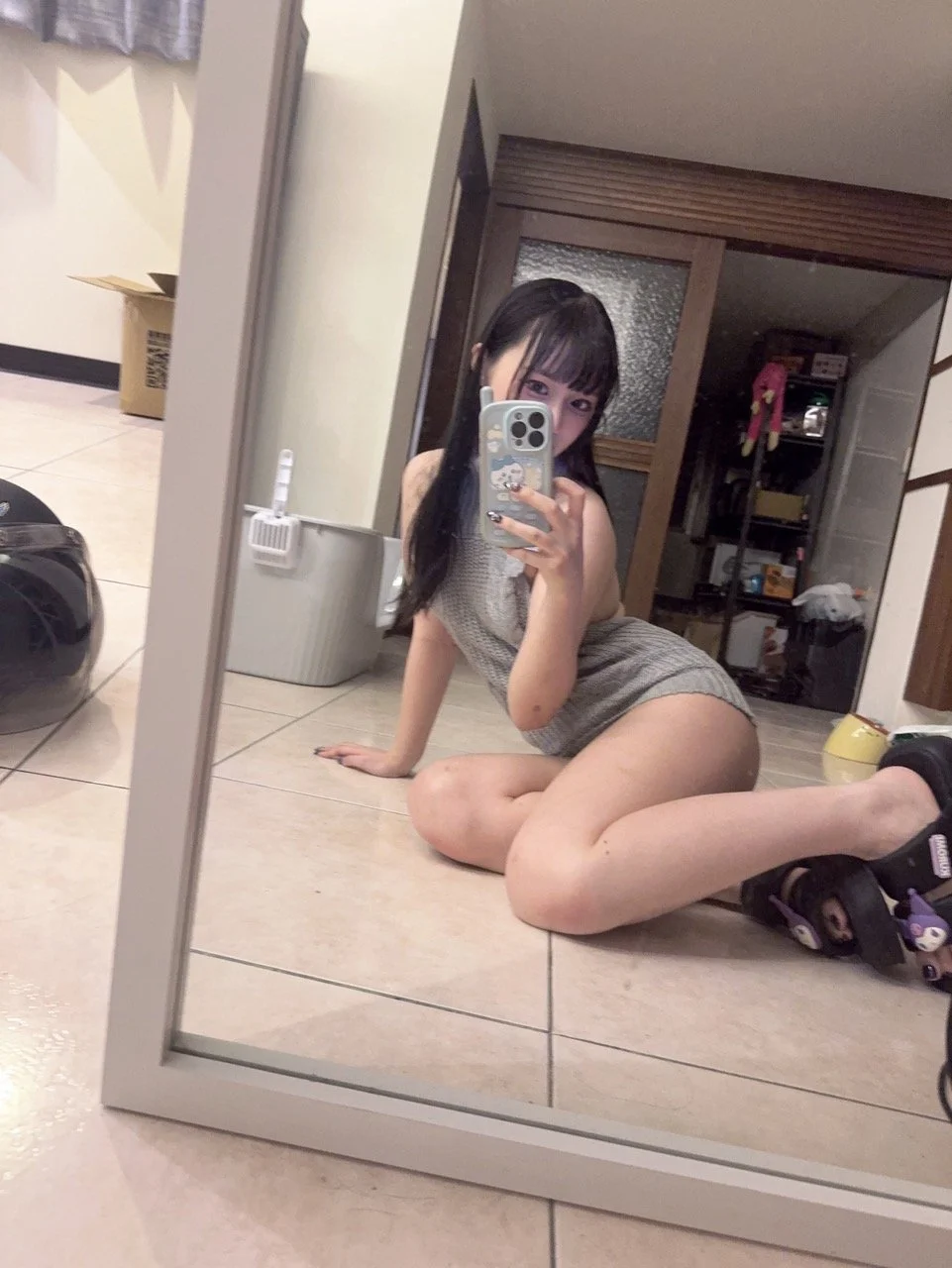 Young woman with long dark hair taking a mirror selfie on the floor inside a room. She is wearing a sleeveless top and shorts, with a phone decorated with cartoon stickers. The room has various objects including shelving and cardboard boxes.