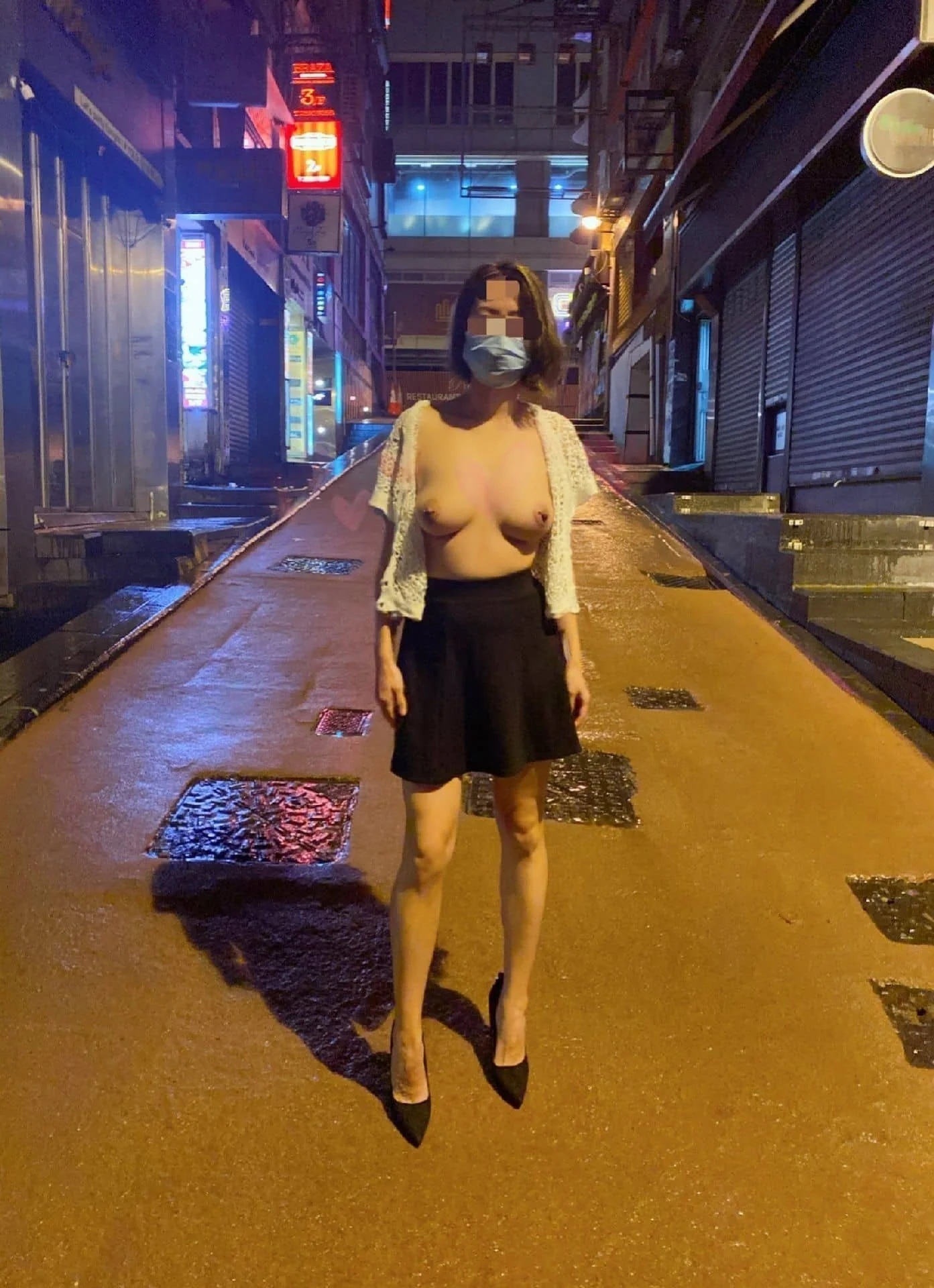 A topless woman wearing a mask, a black skirt, high heels, and a crocheted cardigan standing on a wet city street at night with illuminated signs and closed shops.