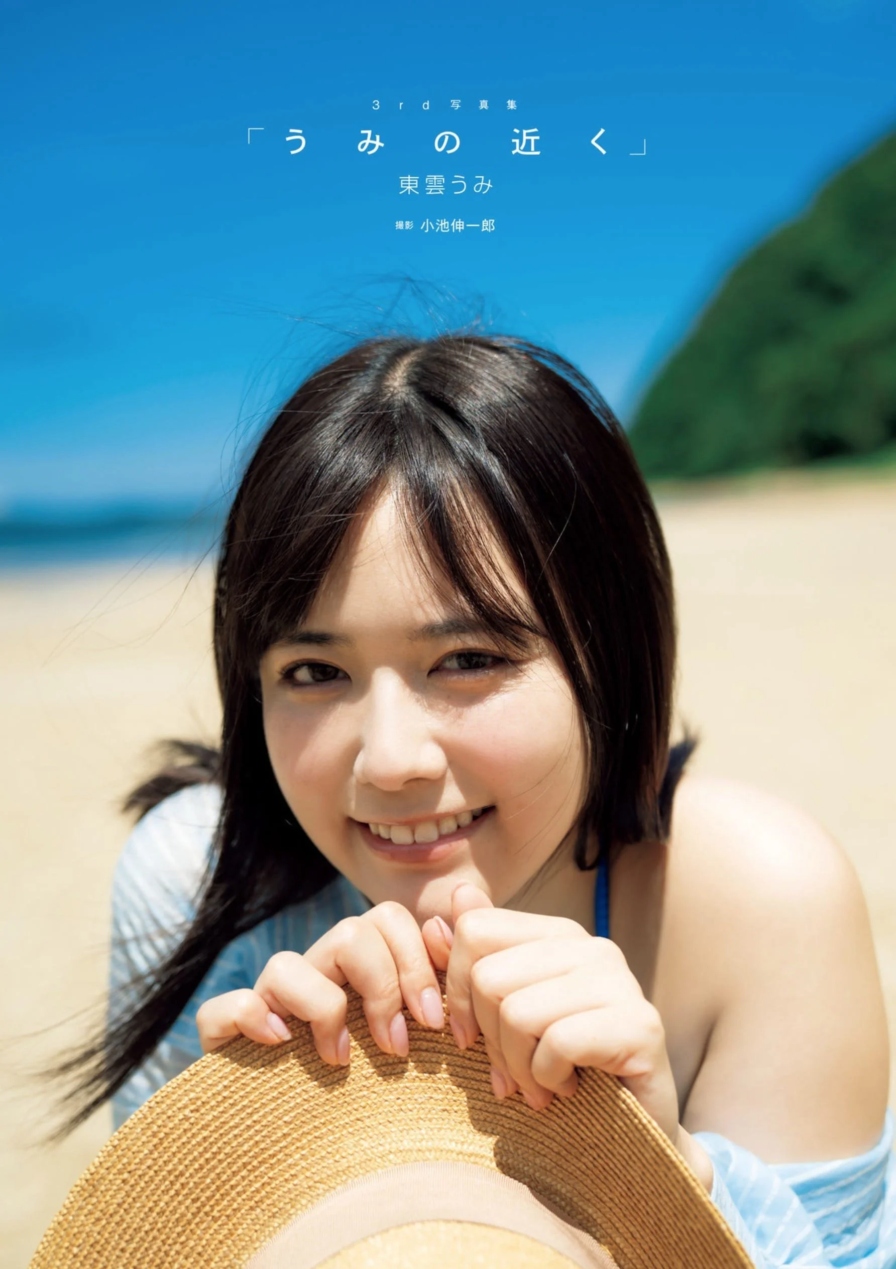 Young woman smiling and holding a straw hat on a sandy beach with green hills and blue sky in the background.