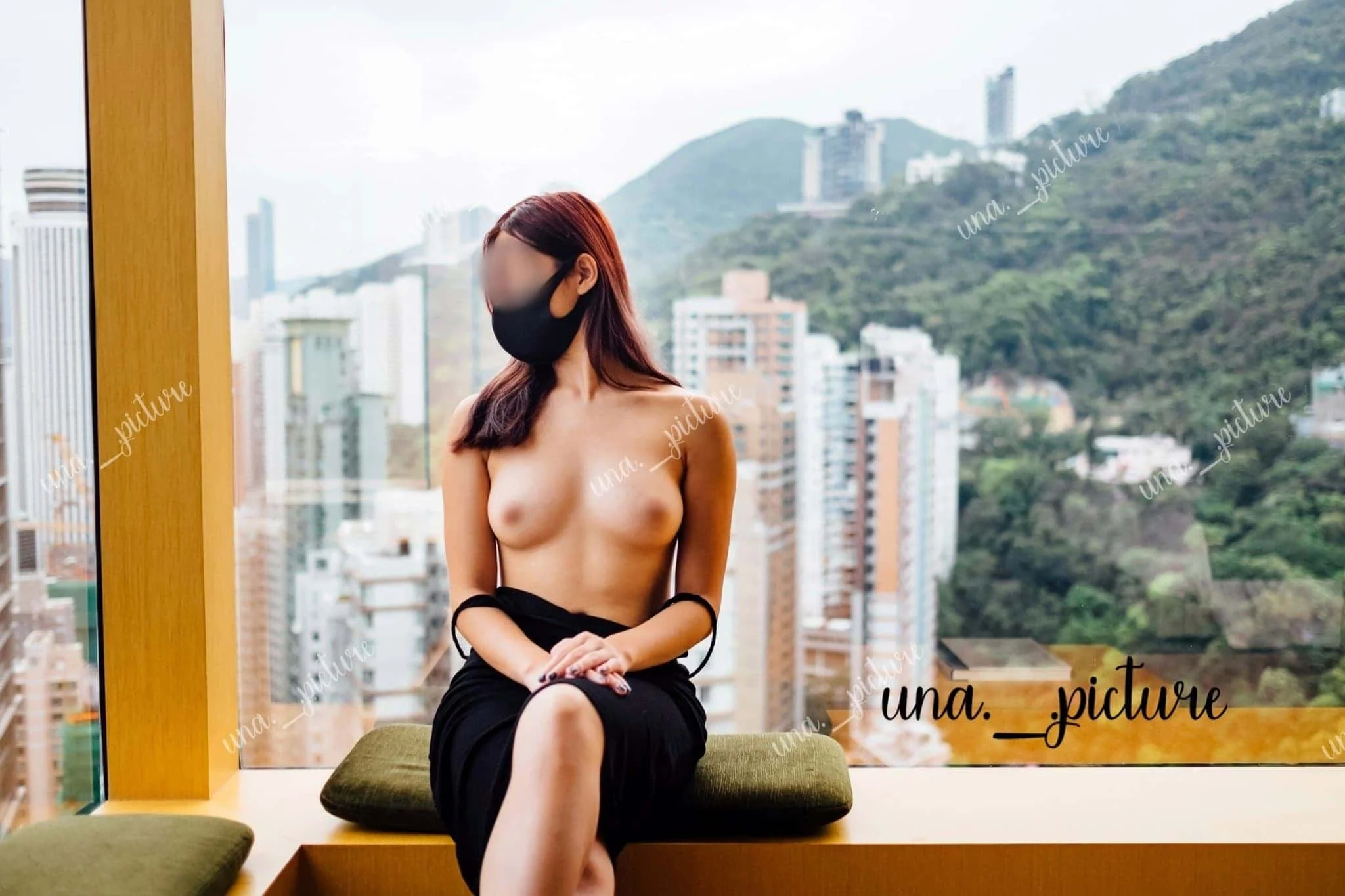 A topless woman wearing a black mask, sitting by a large window with a cityscape and green mountain in the background.
