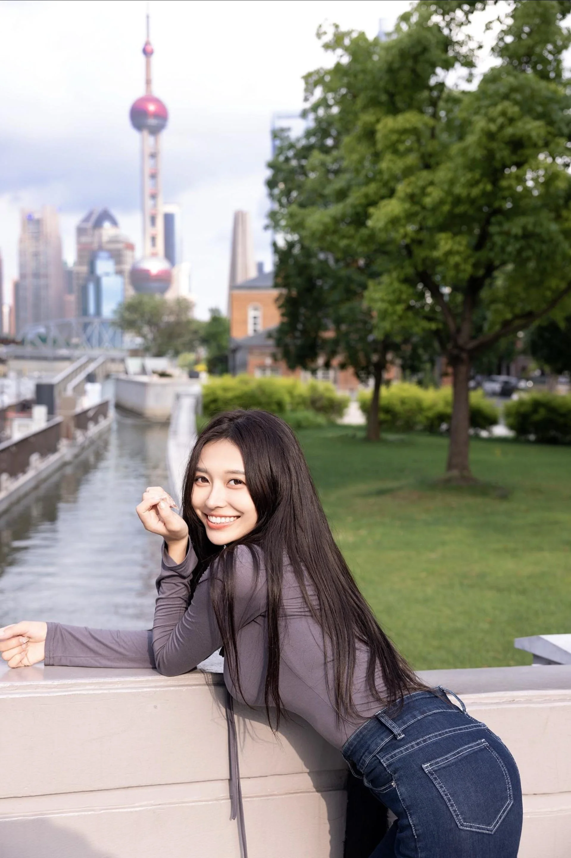 Young woman with long dark hair leaning on a bridge railing near a waterway in an urban park, with a city skyline featuring tall buildings and a distinctive tower in the background during daytime.