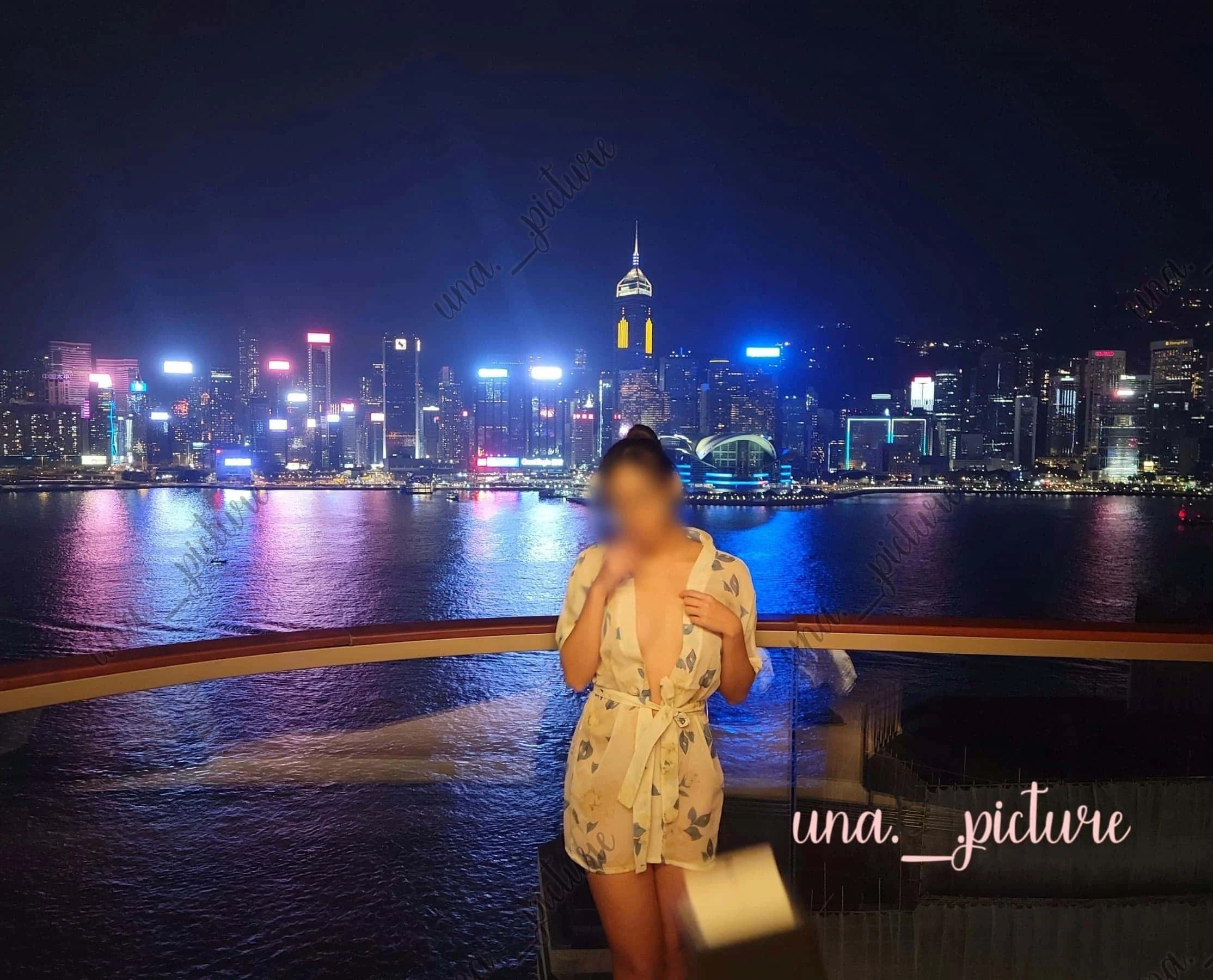 A woman in a floral robe standing on a balcony overlooking a brightly lit city skyline at night with water in the foreground.