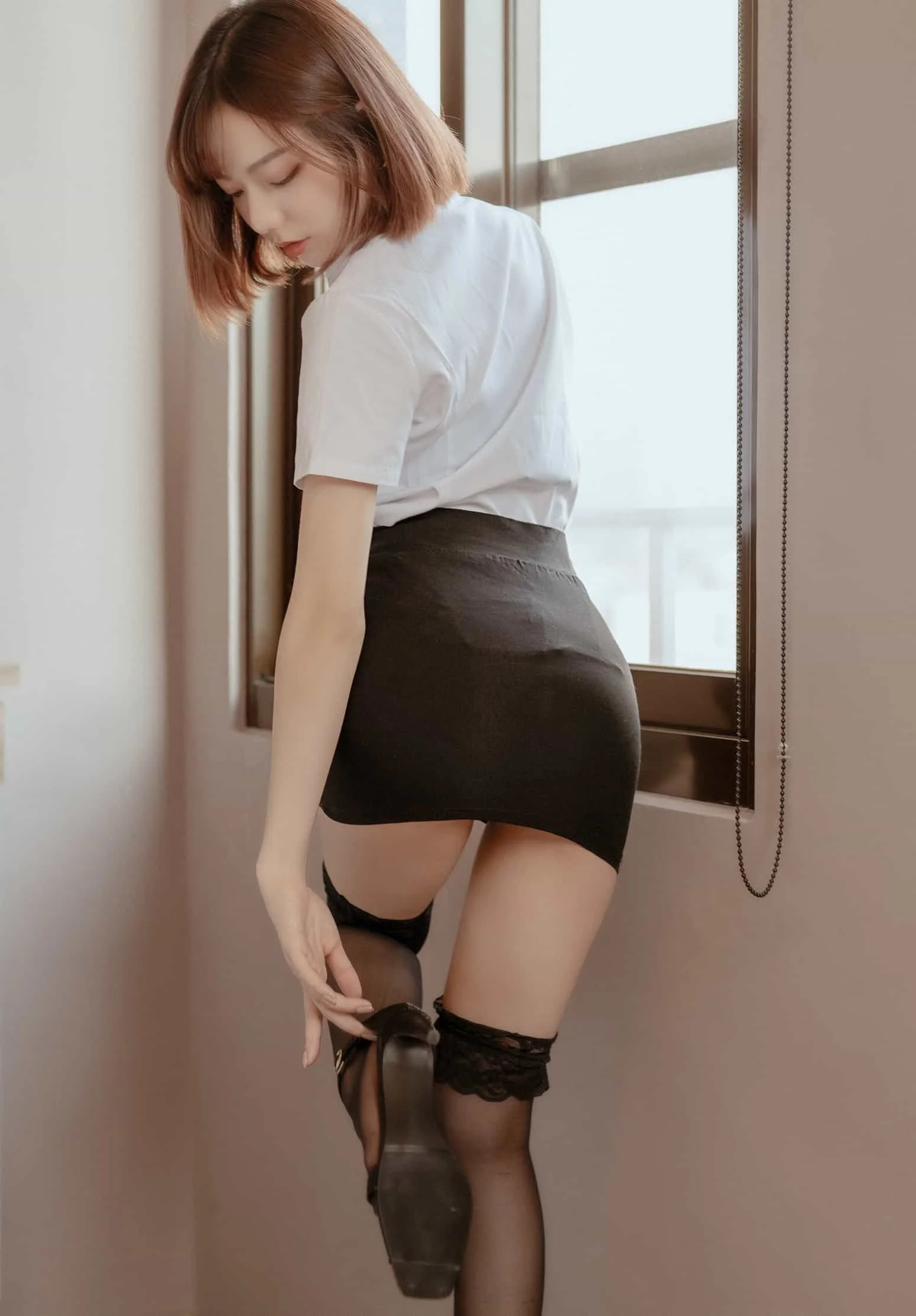 Person in white shirt and black skirt near window, adjusting black stockings.