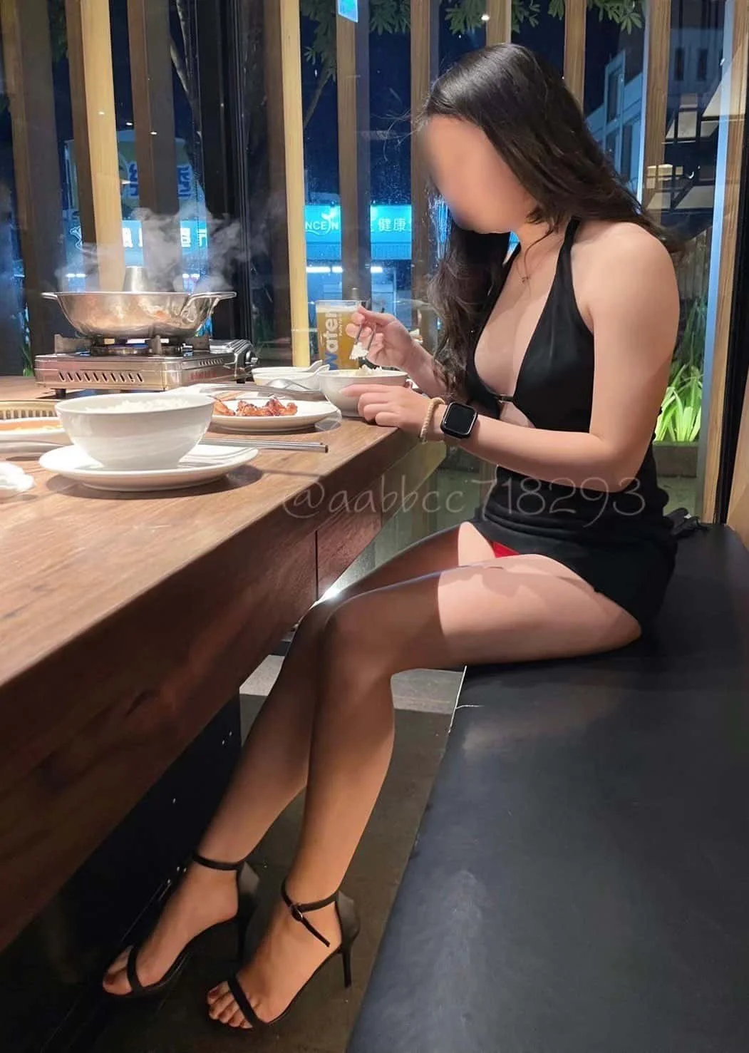 A woman with long dark hair sits at a restaurant table, wearing black high heels, a black top, and shorts, eating a meal with steam rising from a hot pot on the table.