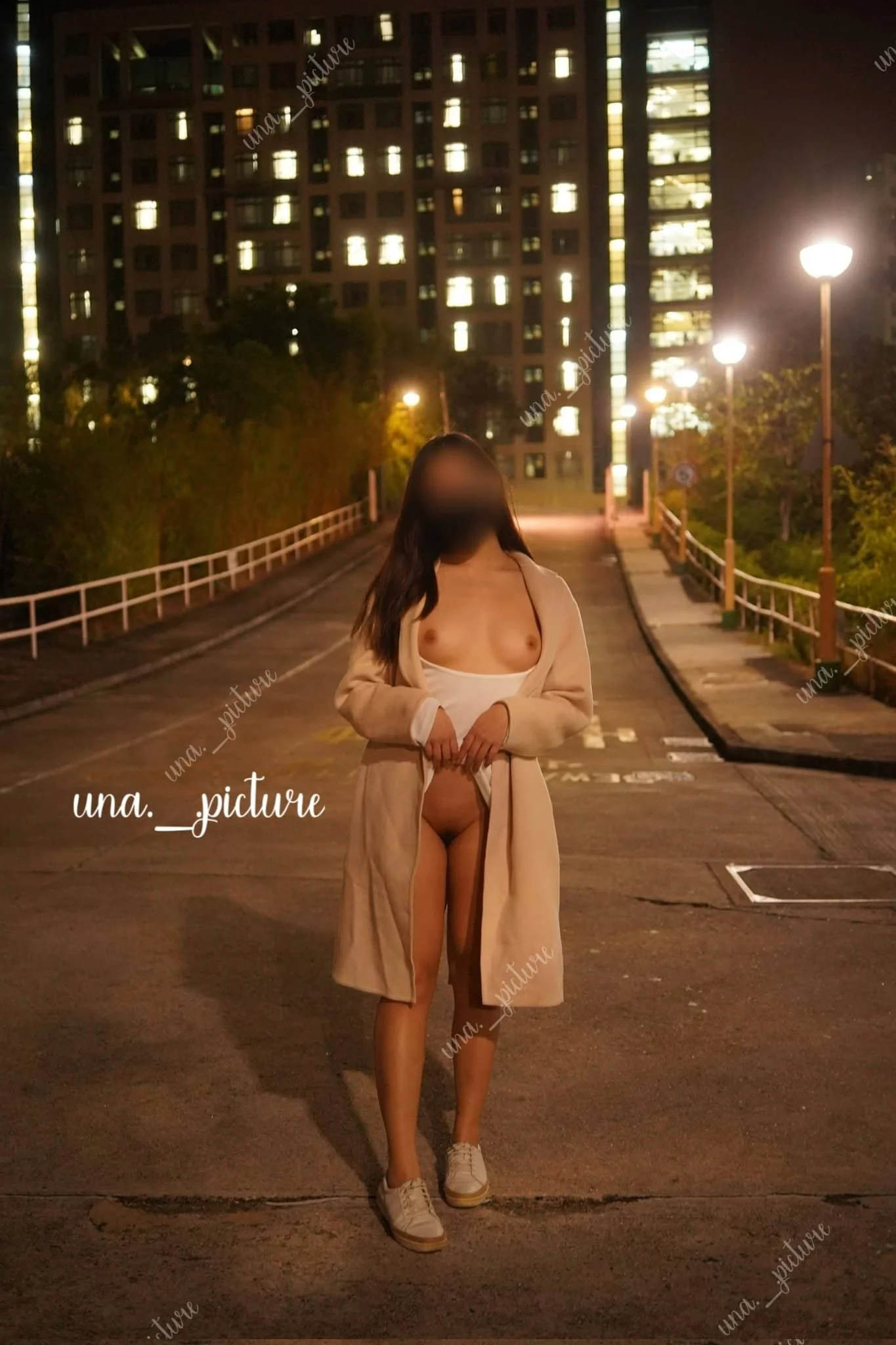 A woman standing topless on a city street at night, wearing a beige coat, white underwear, and sneakers, with a backdrop of tall buildings and streetlights.