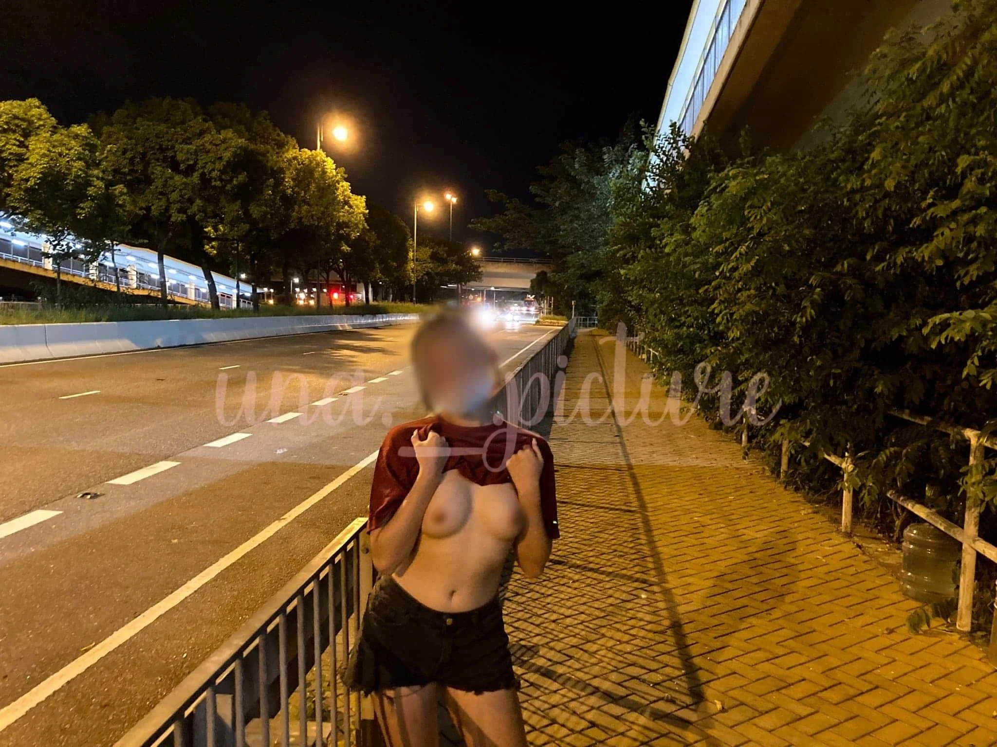 A woman with blurred face standing on a city sidewalk at night, lifting her shirt to reveal her bare chest.