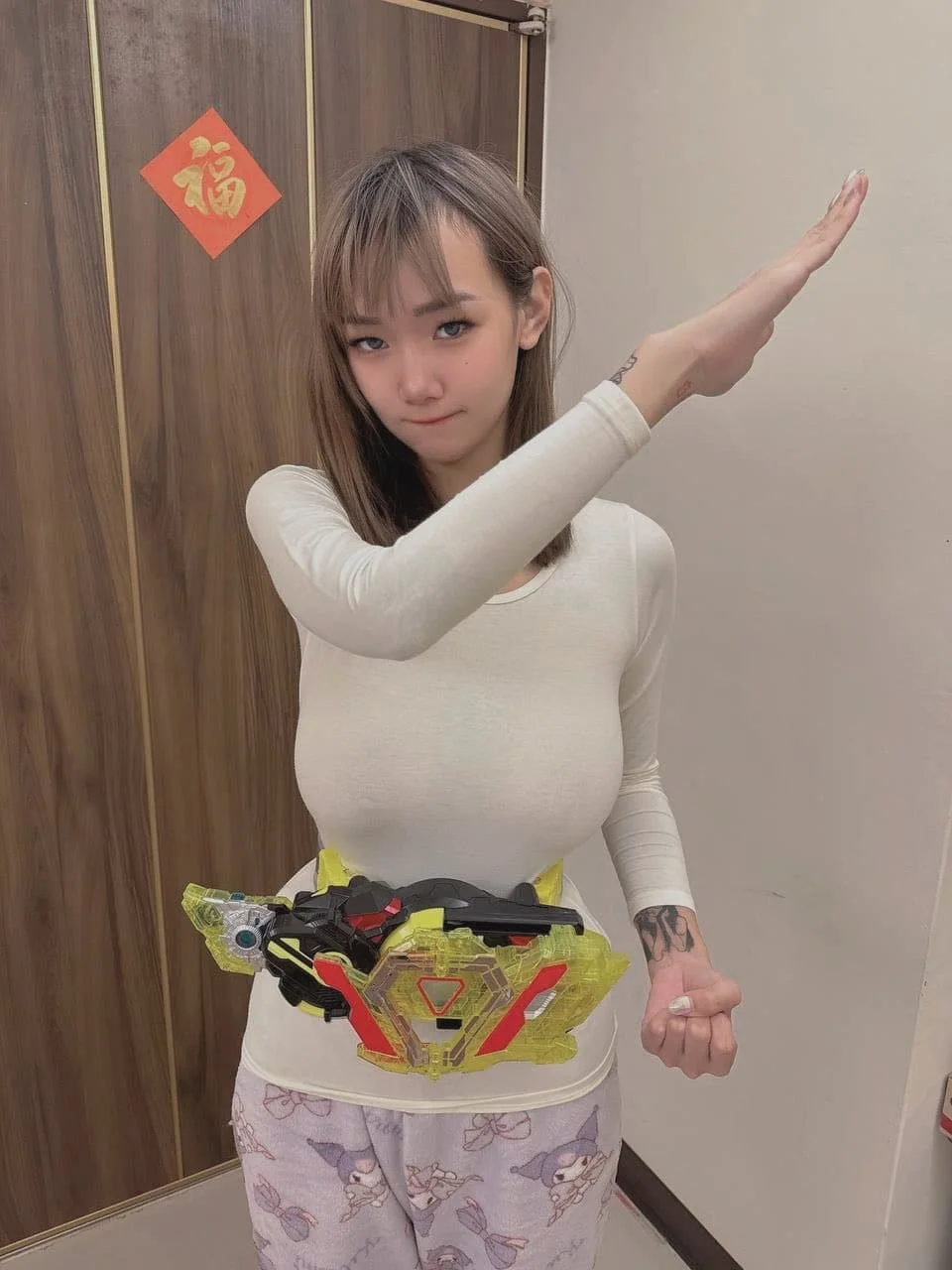 A young woman with light brown hair and bangs is posing with her right arm raised and her left arm bent into a fist, wearing a white long-sleeve shirt and pajama pants with a cartoon character print. She has a belt with a colorful plastic toy weapon 