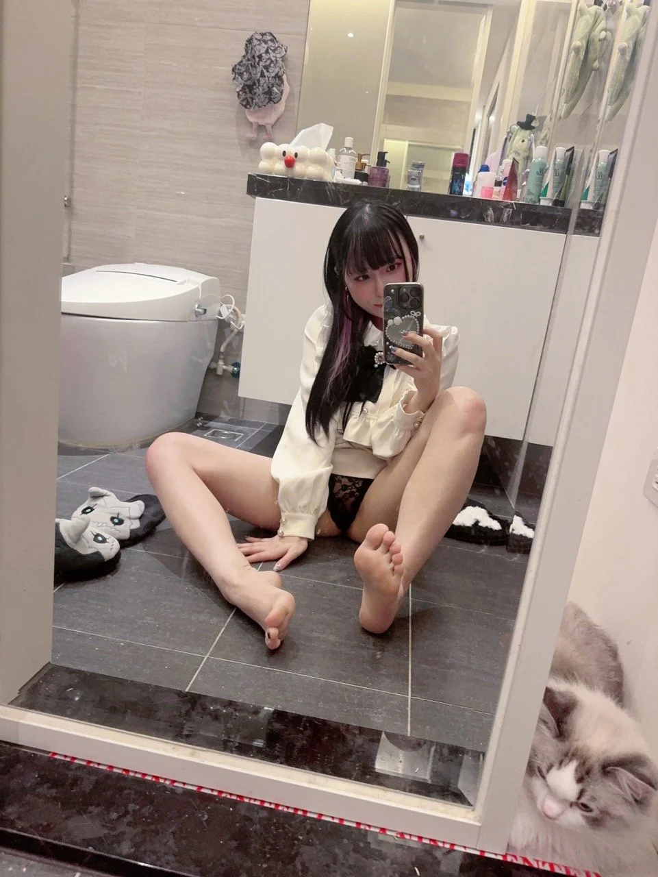 Young woman sitting on bathroom floor taking a mirror selfie, wearing a cream-colored blouse and black lace underwear, with a cat sitting near the mirror's edge and slippers nearby, bathroom with toiletries and decorations in background.